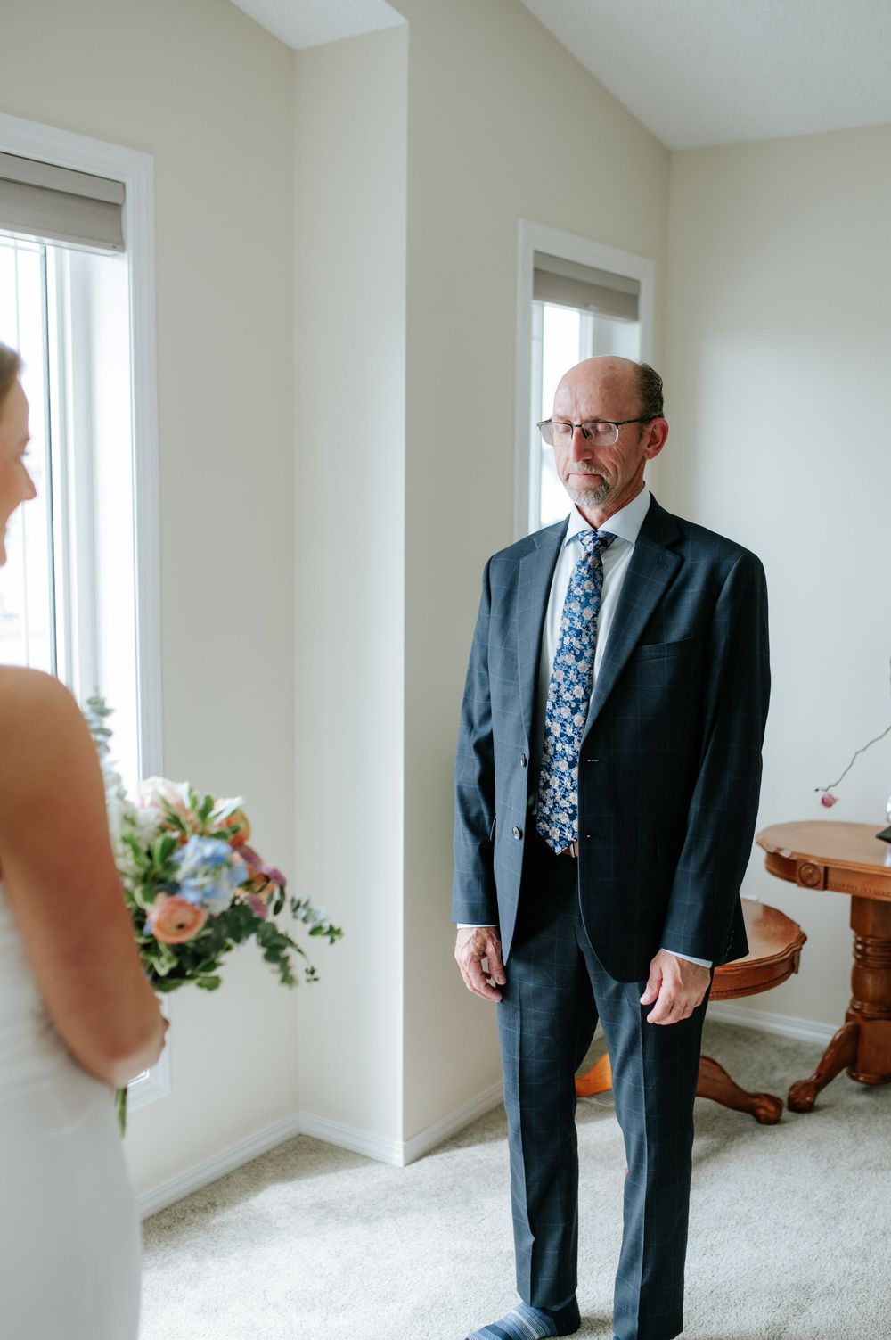 A gentleman in a dark suit and floral tie stands in a bright room with white walls.