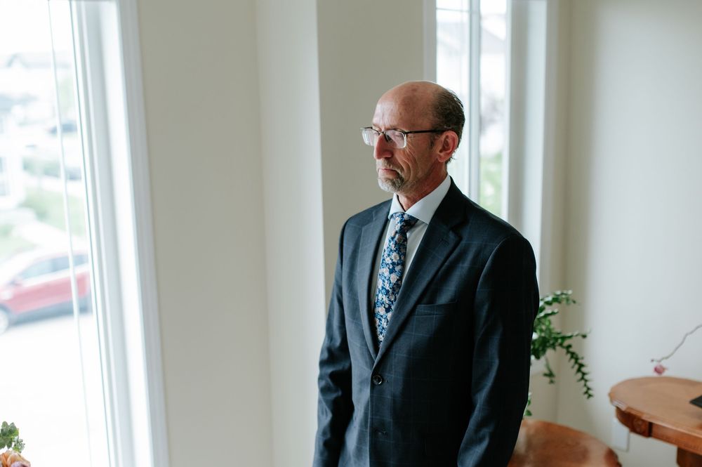A person in a dark business suit and tie stands near a bright window with natural light streaming in.