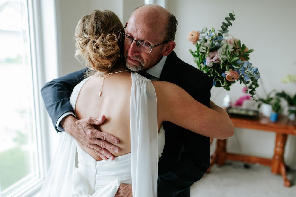 A tender embrace between a bride and groom in formal wedding attire near a bright window.