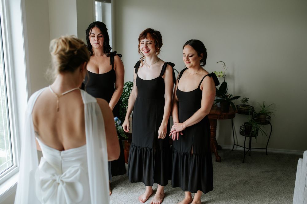 A group of people wearing black dresses gather near a window during a wedding preparation.