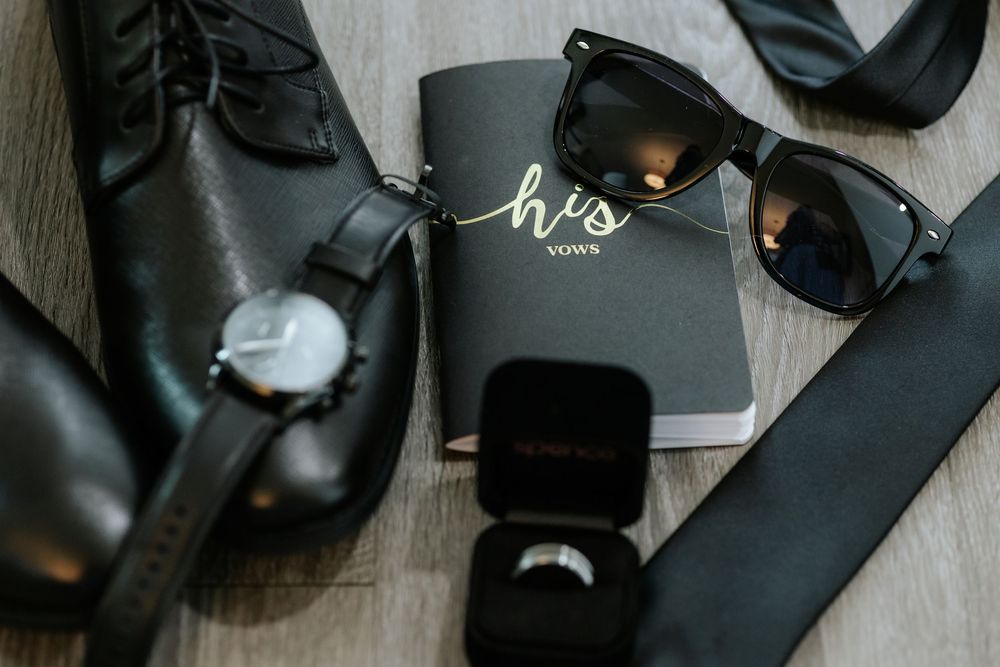 Wedding details including rings, black leather shoes, and accessories laid out on a dark surface.
