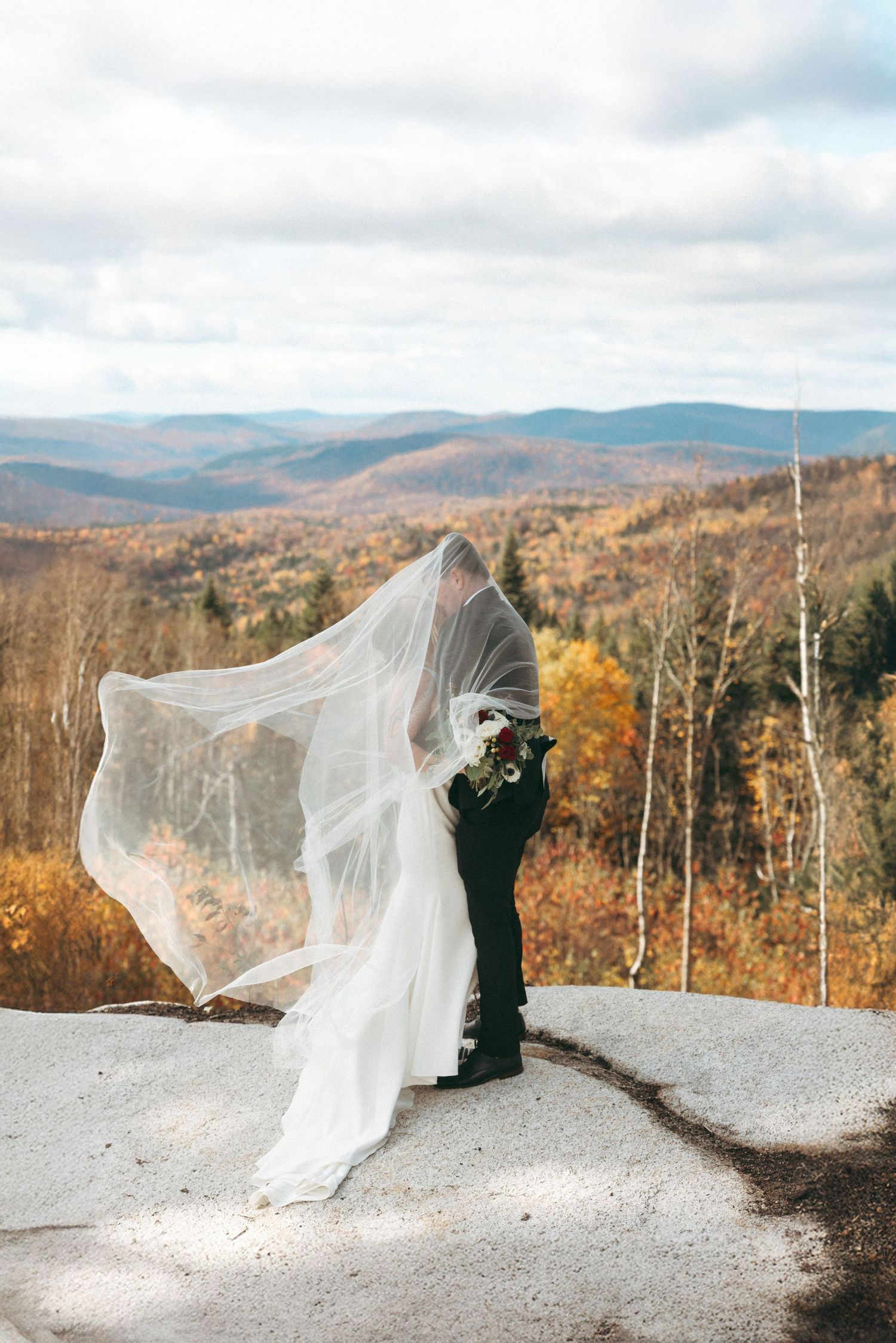 A wedding couple stands on a mountain overlook during autumn with a flowing veil catching the breeze against fall foliage.