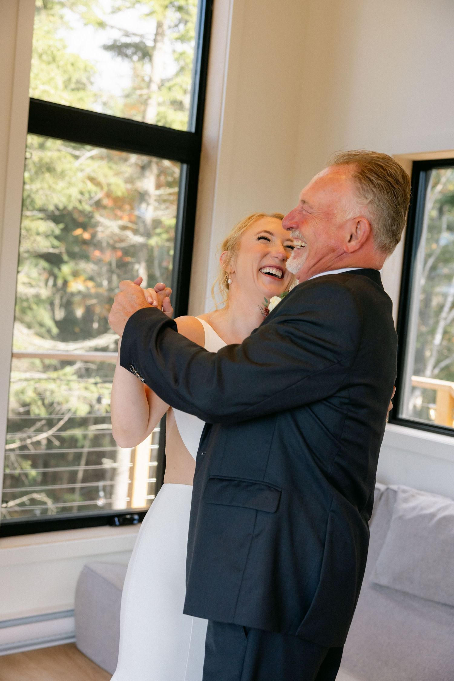A couple in formal attire share a joyful dance moment in a sunlit room.