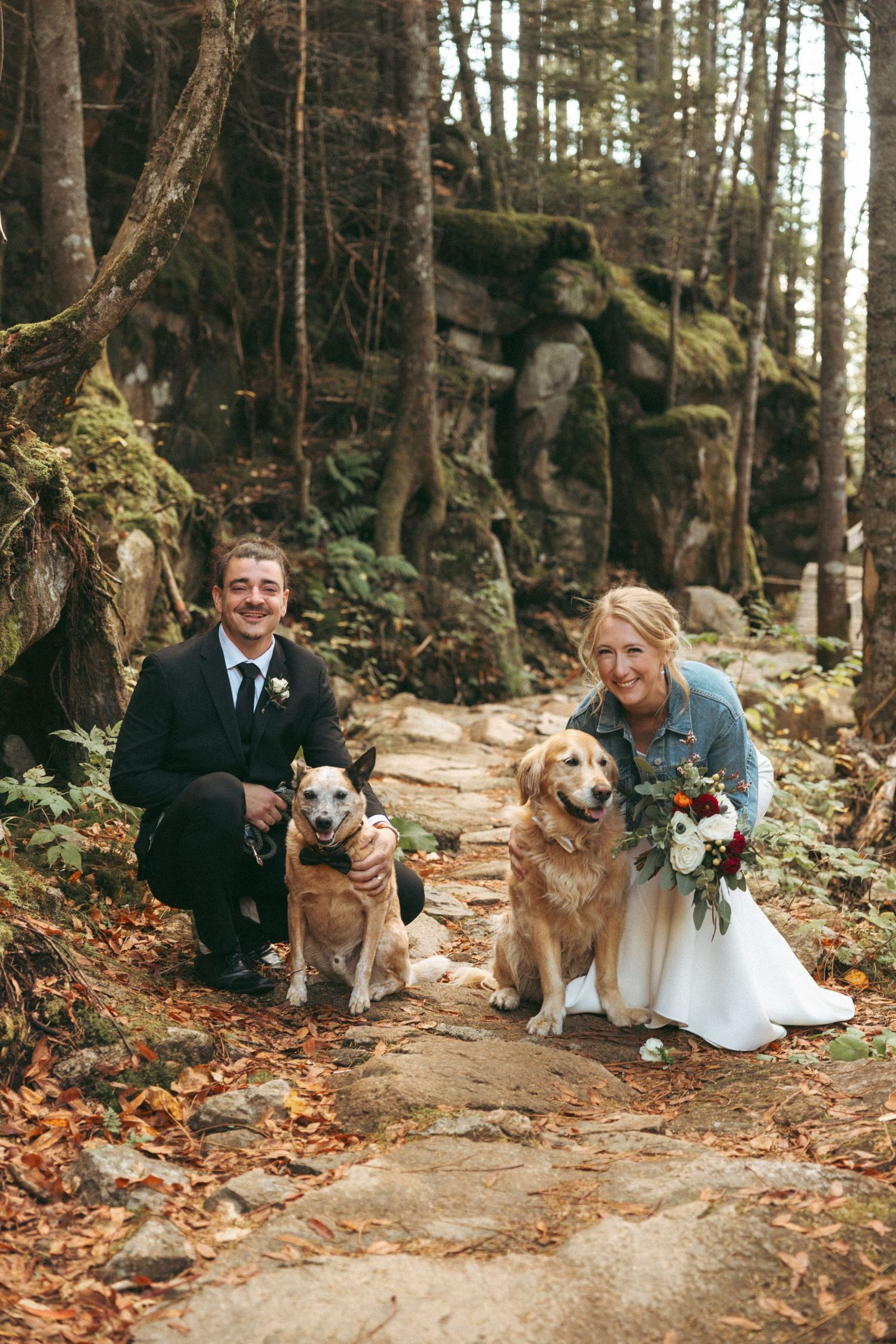 A couple poses with their dog on a forest path covered in autumn leaves during a wedding photoshoot.
