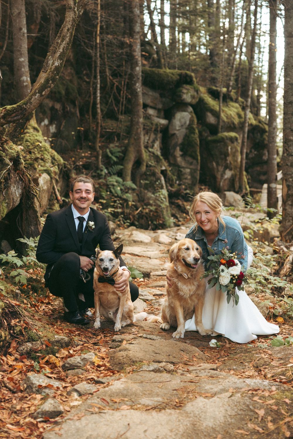 A couple poses with their dog on a forest path covered in autumn leaves during a wedding photoshoot.