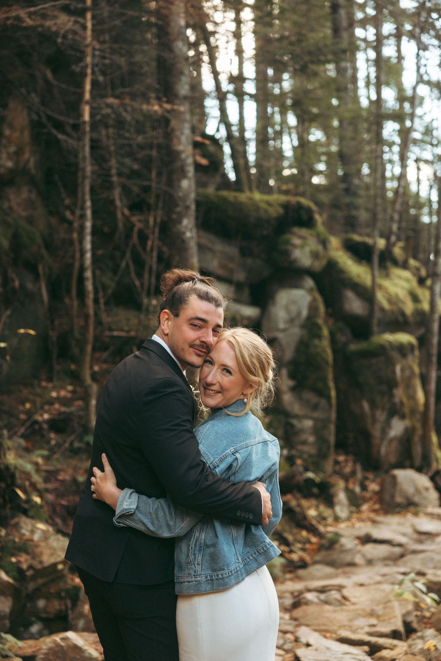 A couple shares a romantic embrace while standing on a rocky forest trail surrounded by tall trees.
