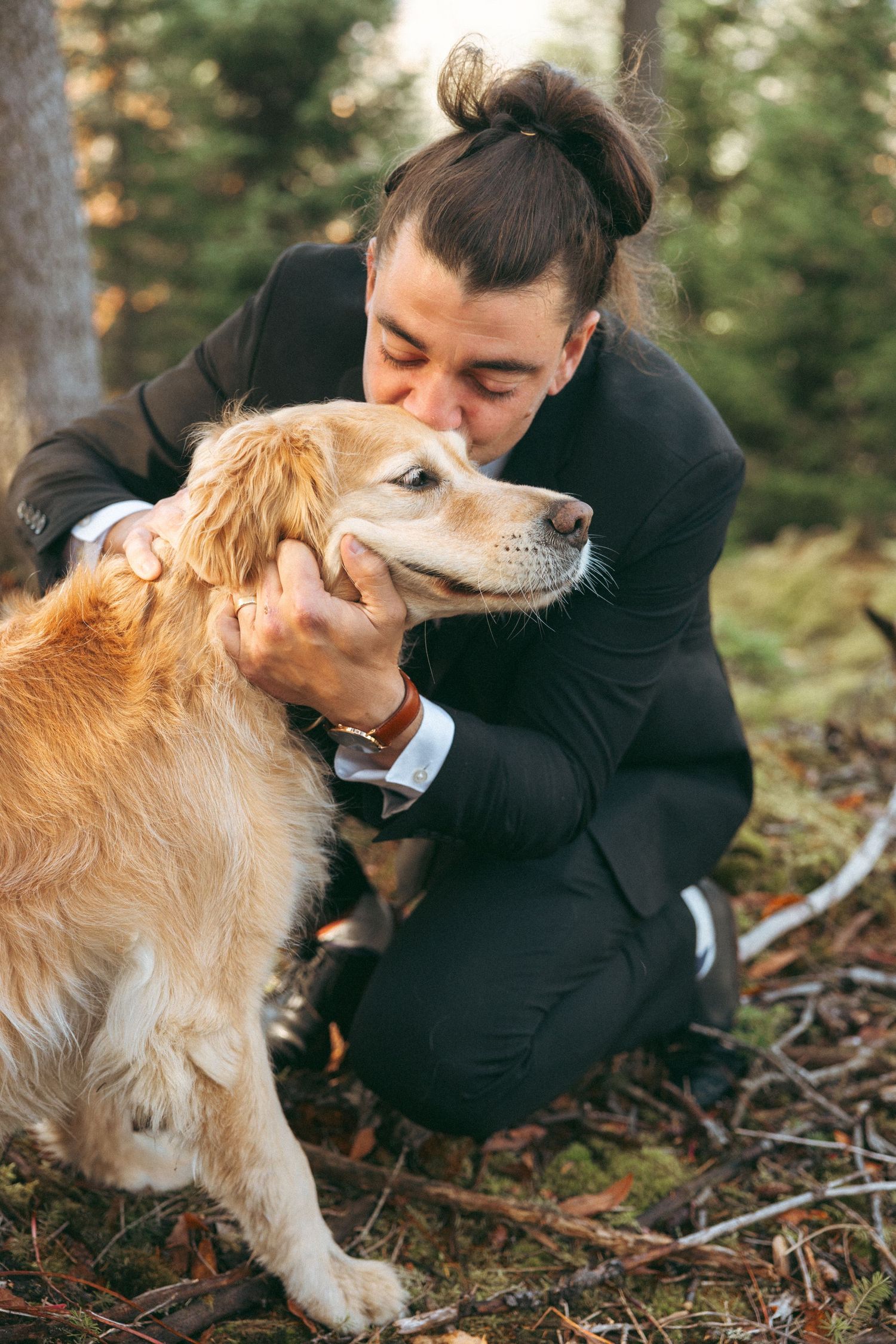 A tender moment between a person in black clothing and a golden retriever dog on a forest floor.