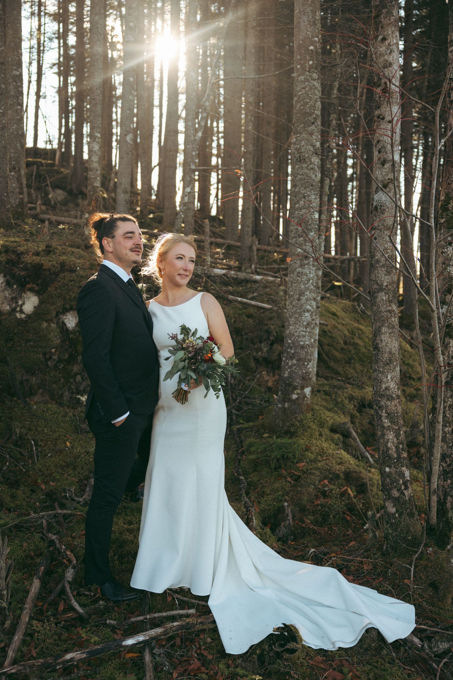 A couple shares a joyful moment in a sunlit forest, the bride wearing a flowing white gown with a long train.