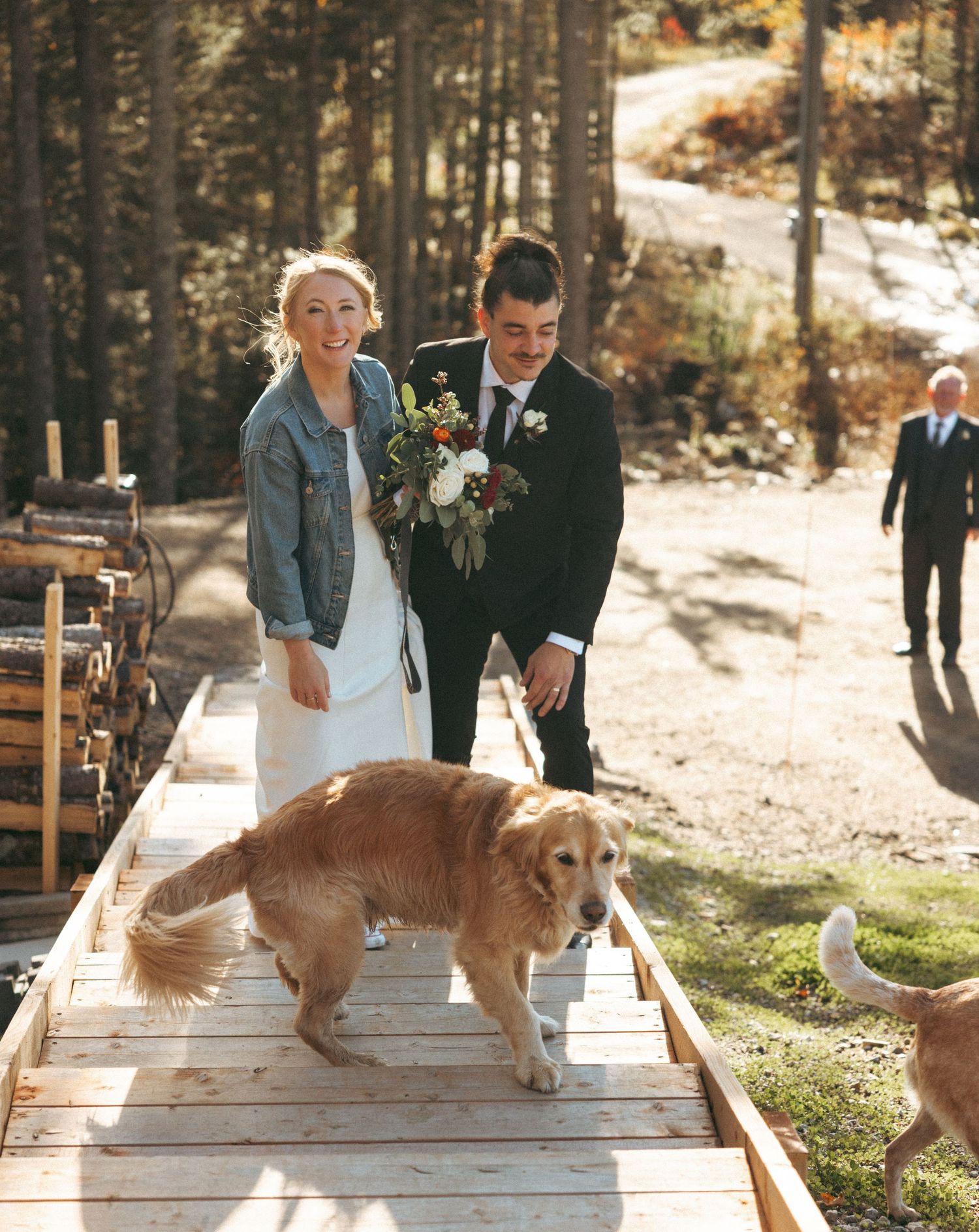 A couple celebrates their outdoor wedding on a wooden walkway with their golden retriever and another dog in a forest setting.