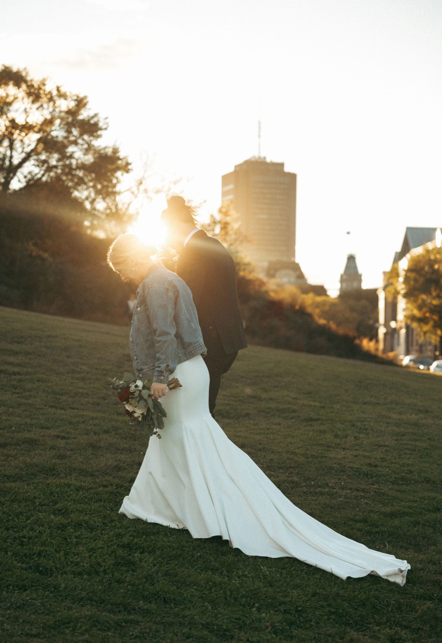 A couple in wedding attire embraces on a grassy hill during golden hour with a cityscape silhouetted in the background.