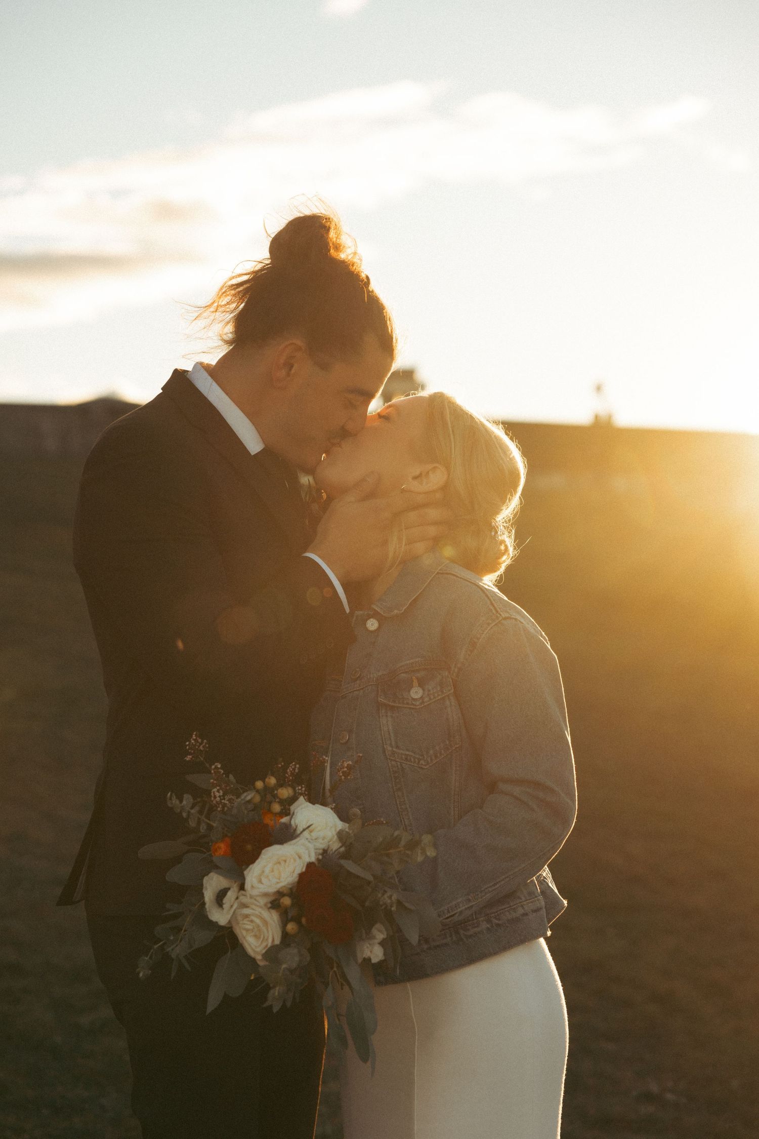 A couple embraces in golden sunset light while holding a white floral bouquet during their intimate wedding moment.