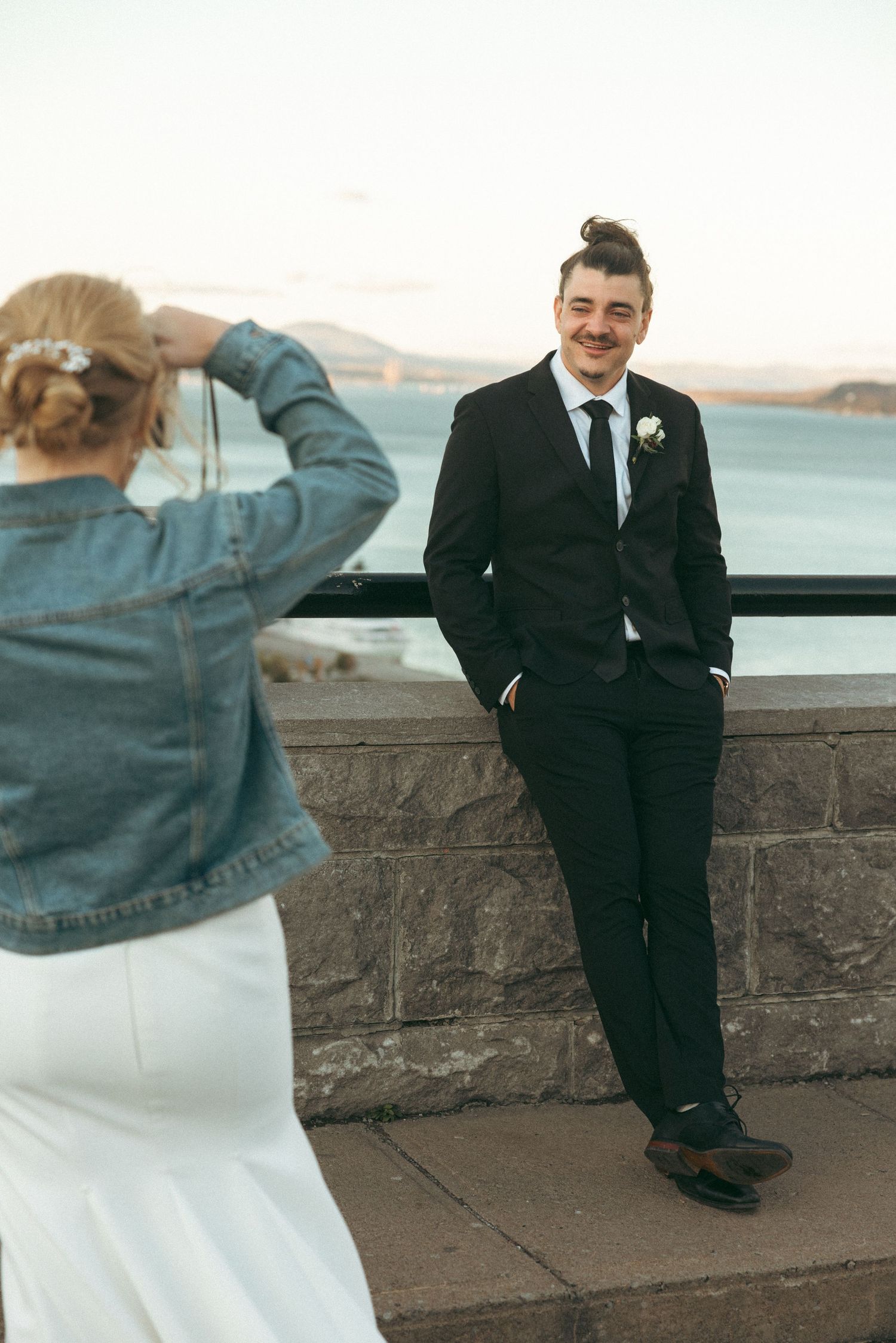 Couple in formal attire poses against stone wall overlooking waterfront on cloudy day.