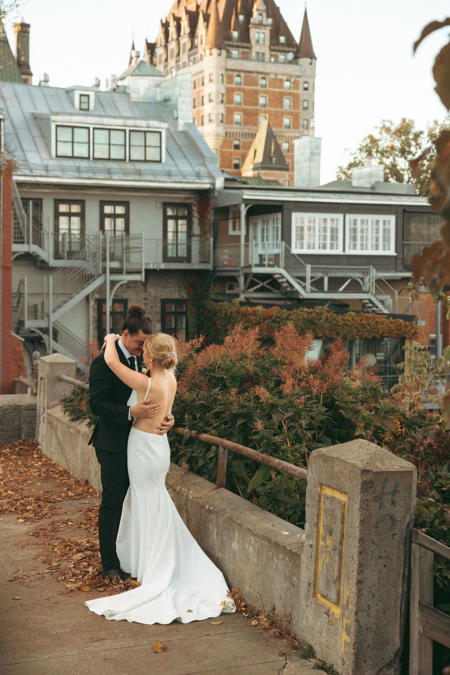 Newlyweds embrace against stone wall with historic architecture and autumn foliage in background.