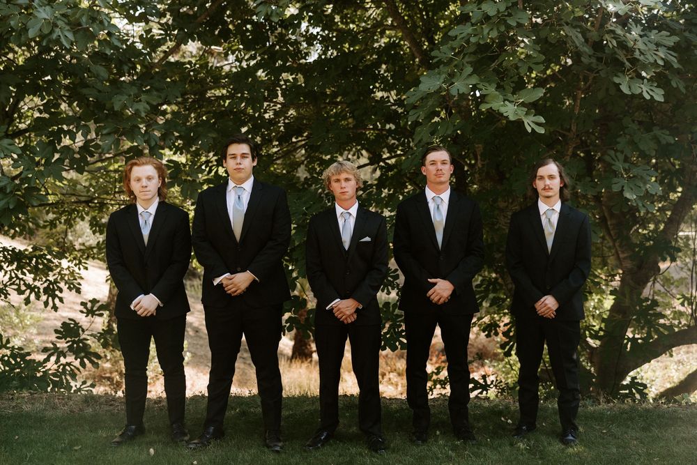 A wedding party in black suits stands in a line outdoors with trees and natural lighting in the background.
