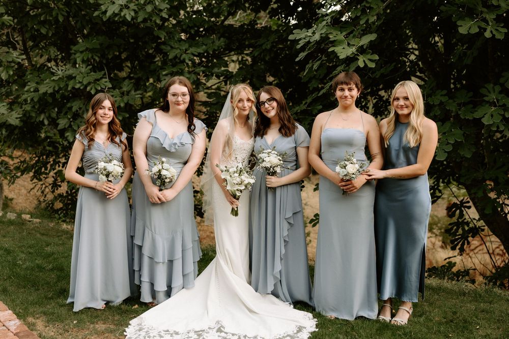 Wedding party in light blue bridesmaid dresses pose together with bride in white gown outdoors in garden setting.