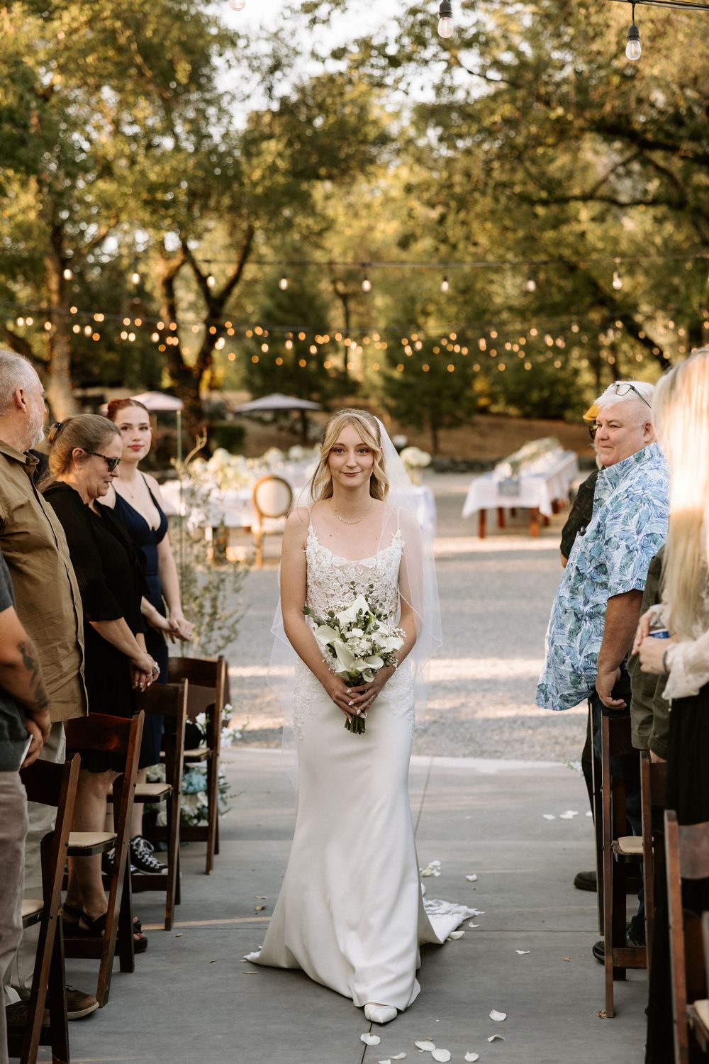 Wedding ceremony setup with wooden chairs and string lights under shady trees.