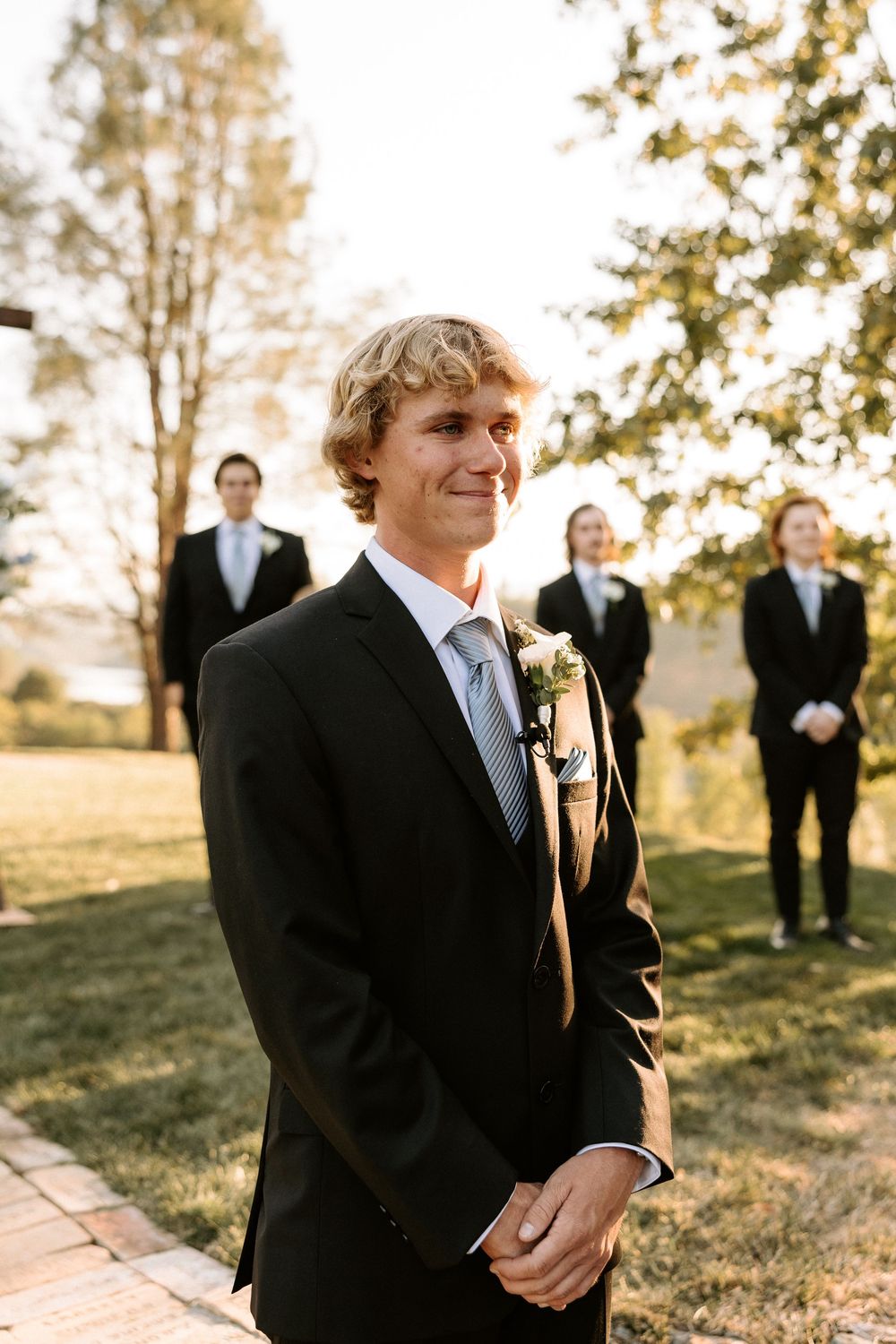 Groomsmen in black suits stand together outdoors during golden hour at a wedding ceremony.