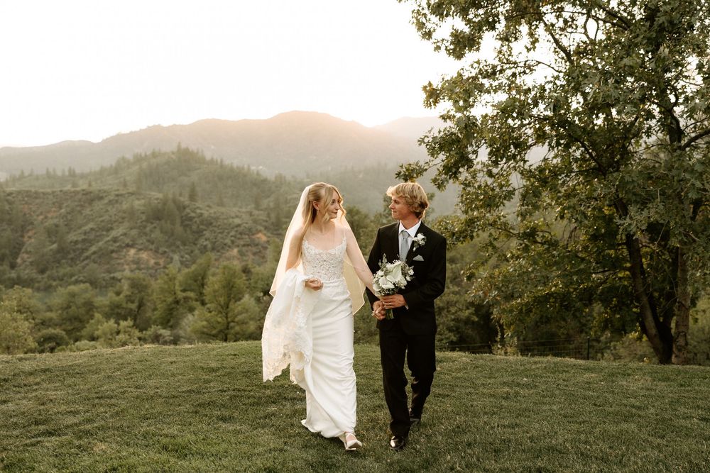 A couple walks hand in hand through a scenic mountain landscape during sunset while dressed in formal wedding attire.