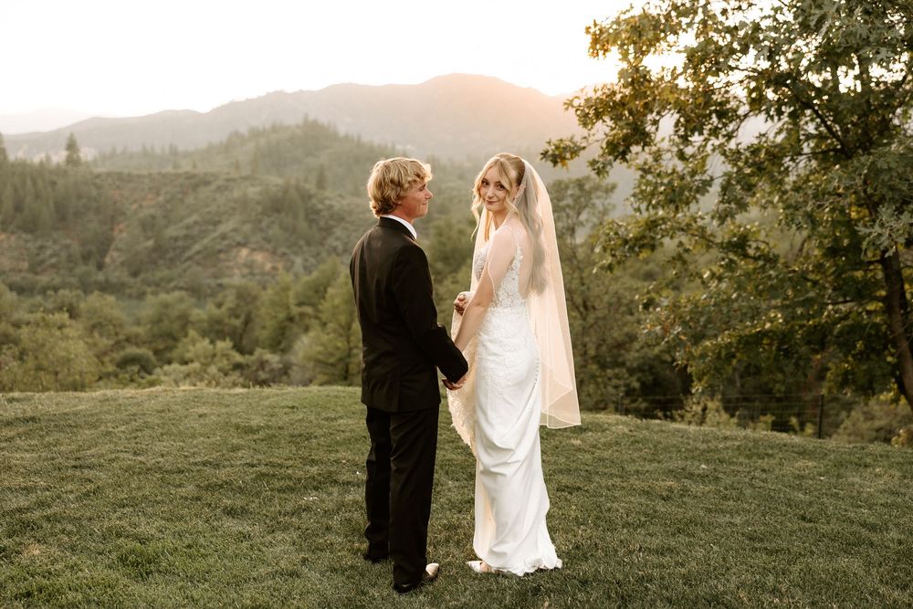 Couple stands together on grassy hilltop during golden sunset in scenic mountain landscape.