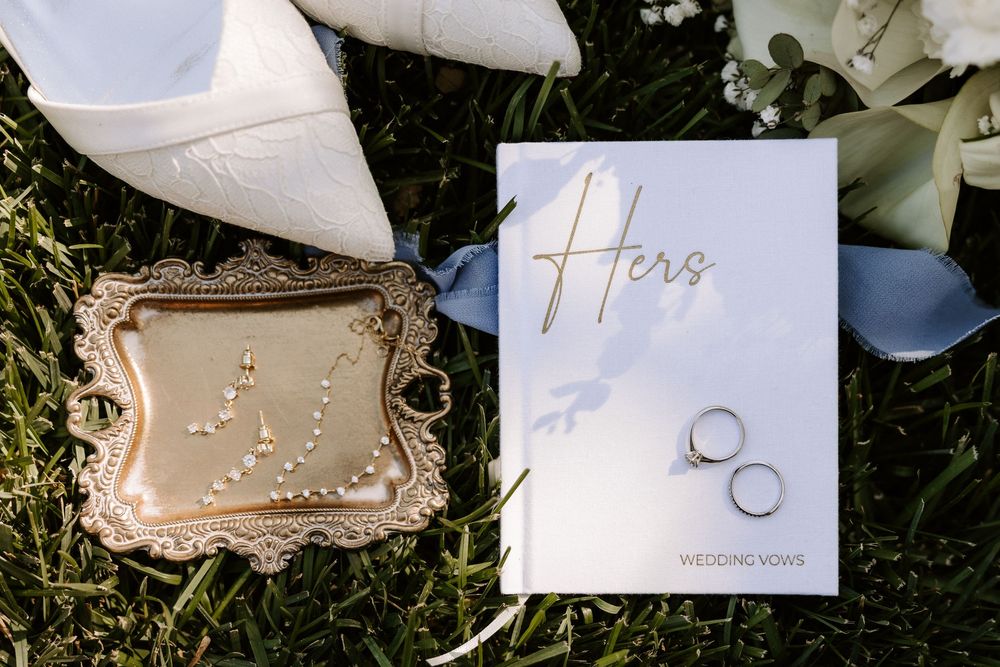 Wedding rings and vows card displayed with a decorative beige purse on grass with floral accents.