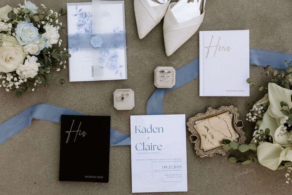 Wedding invitation suite with blue and white stationery, white flowers, and ring box displayed on gray surface.