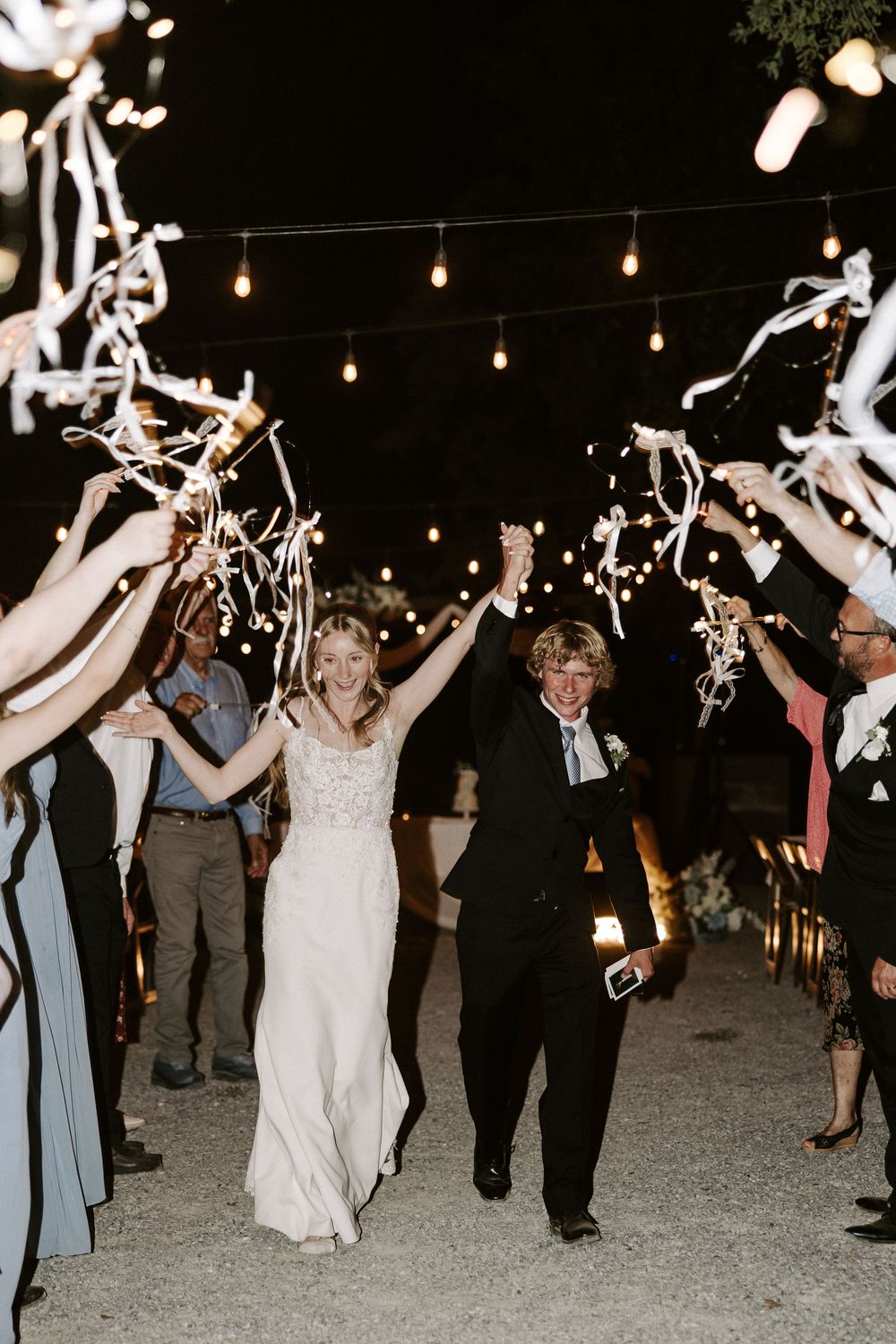 Wedding party celebrates at night under string lights while waving white streamers during a festive outdoor reception.