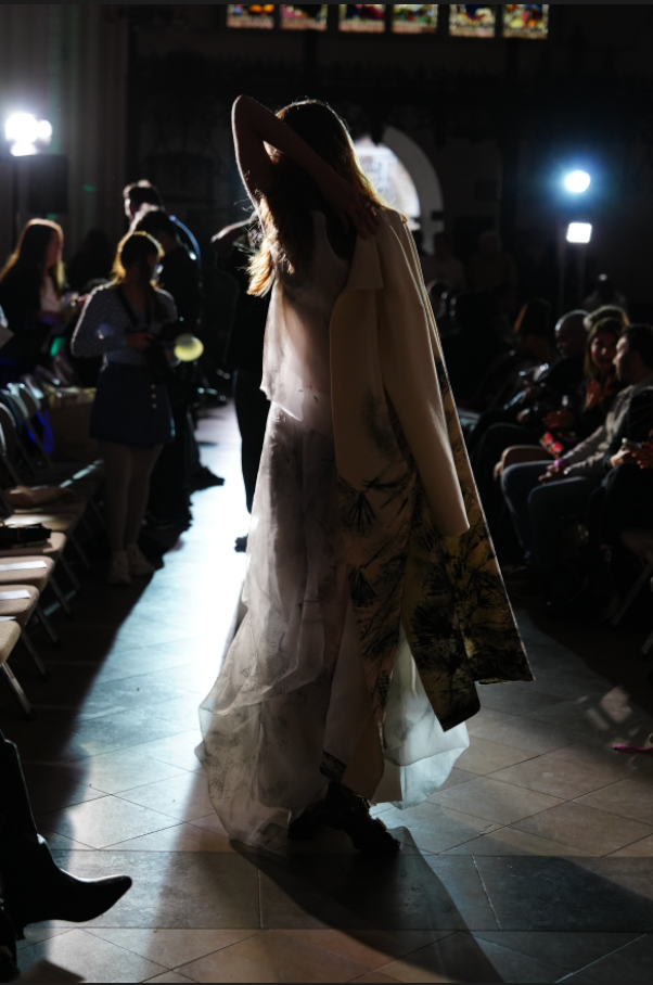 Silhouetted figures and flowing dresses create dramatic shadows on a dark stage during a fashion show performance.