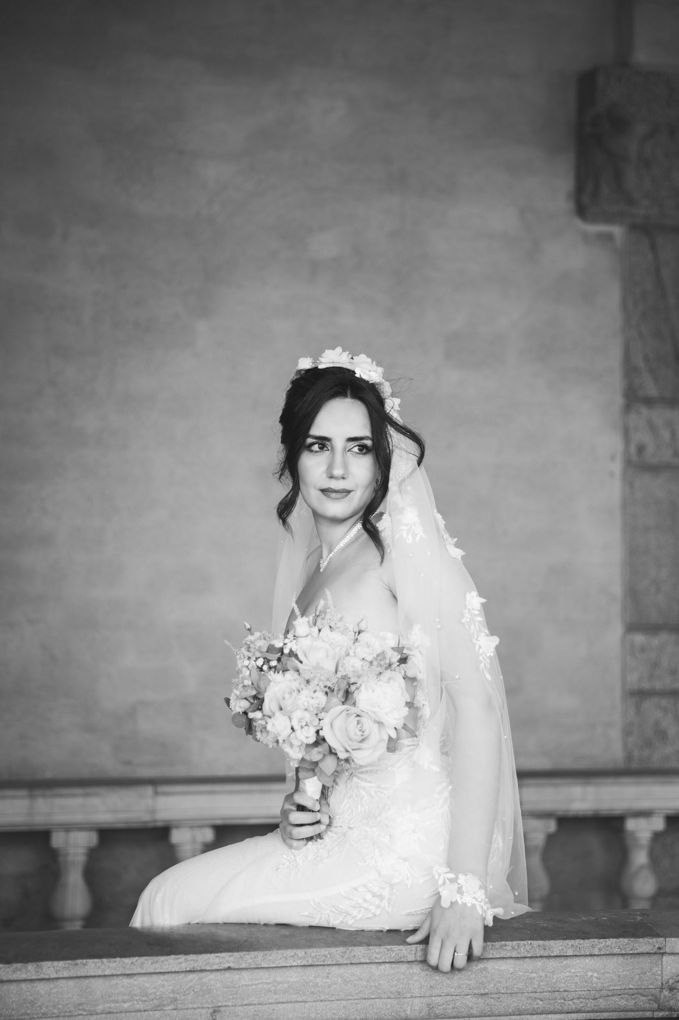 Black and white portrait of a bride in a lace wedding dress and veil holding a floral bouquet while seated.