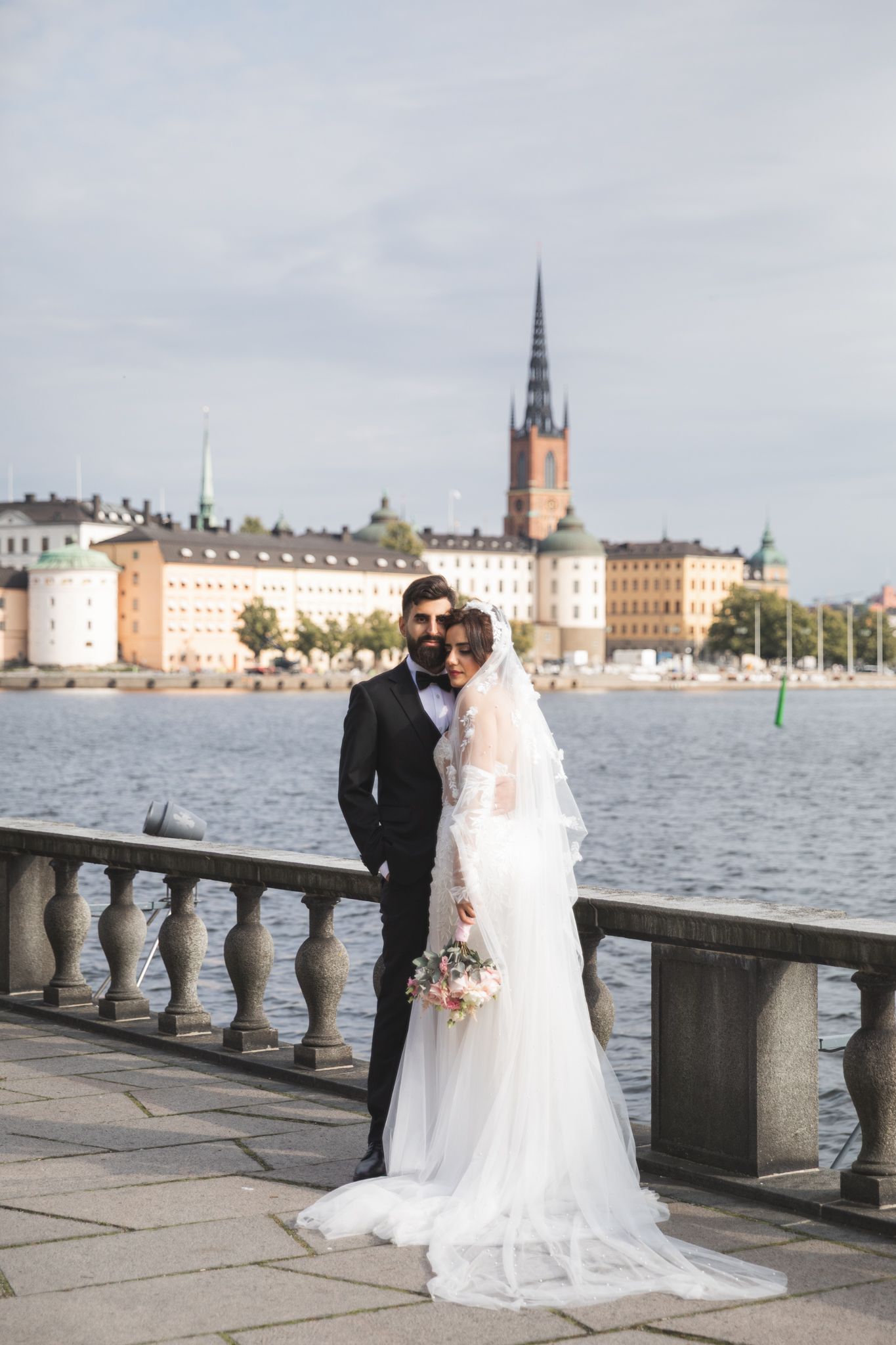 A couple in formal wedding attire poses by a waterfront stone balustrade with historic buildings in the background.