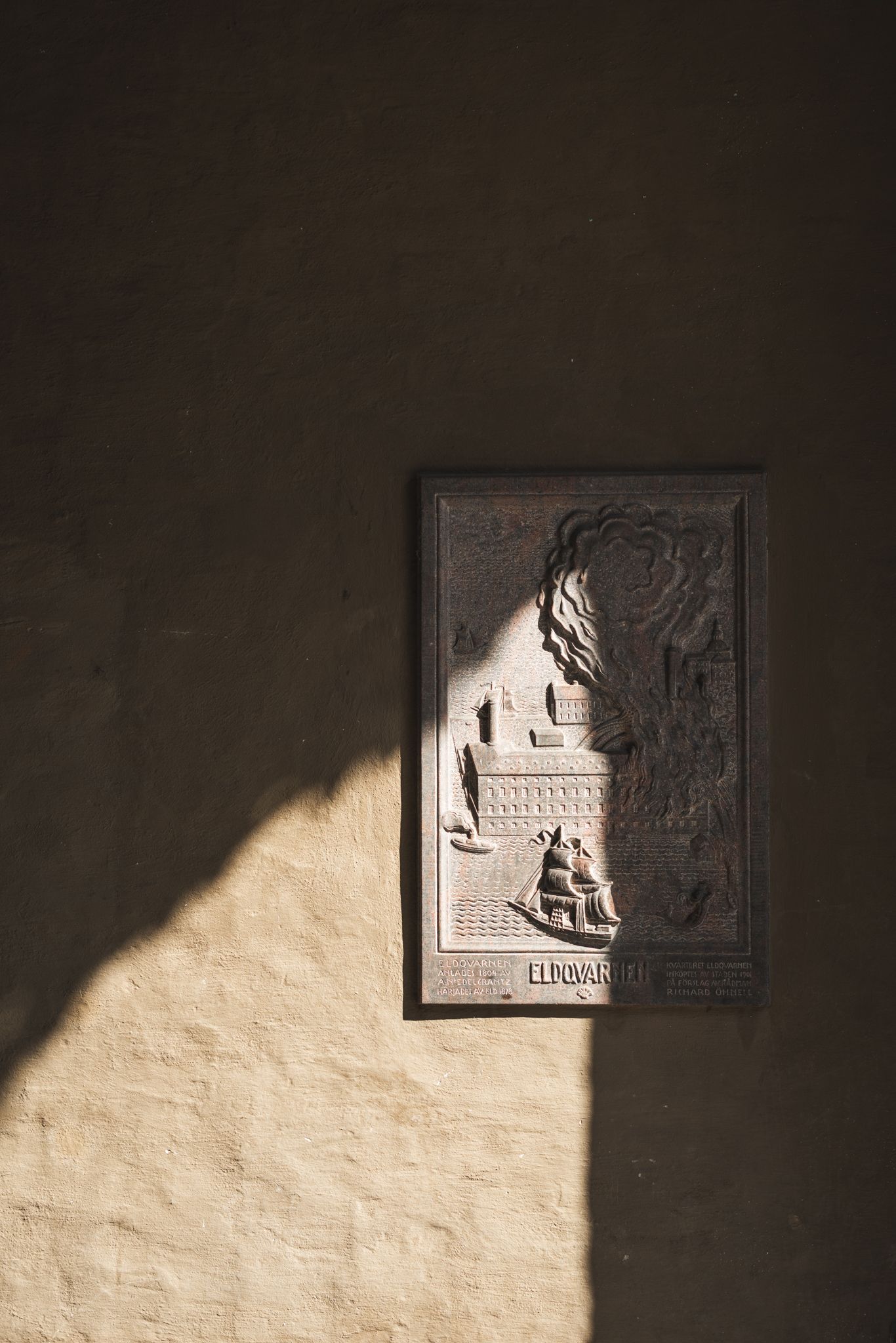 Sunlight casts dramatic shadows on a white wall with mounted decorative stone relief panels.