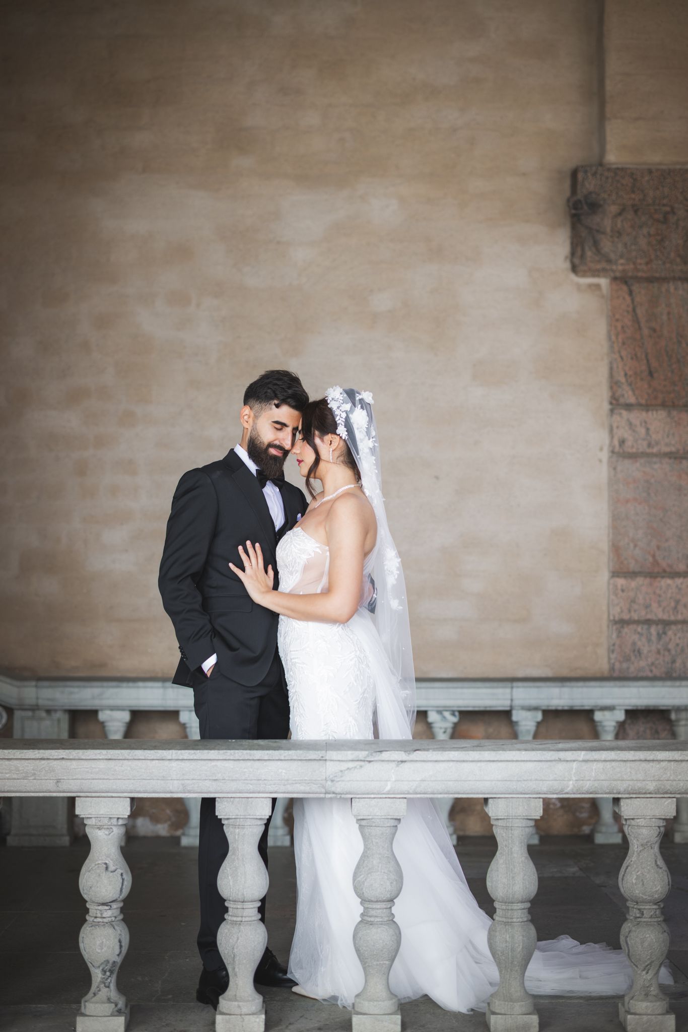 A romantic wedding couple embraces against a stone balustrade wall in an elegant architectural setting.