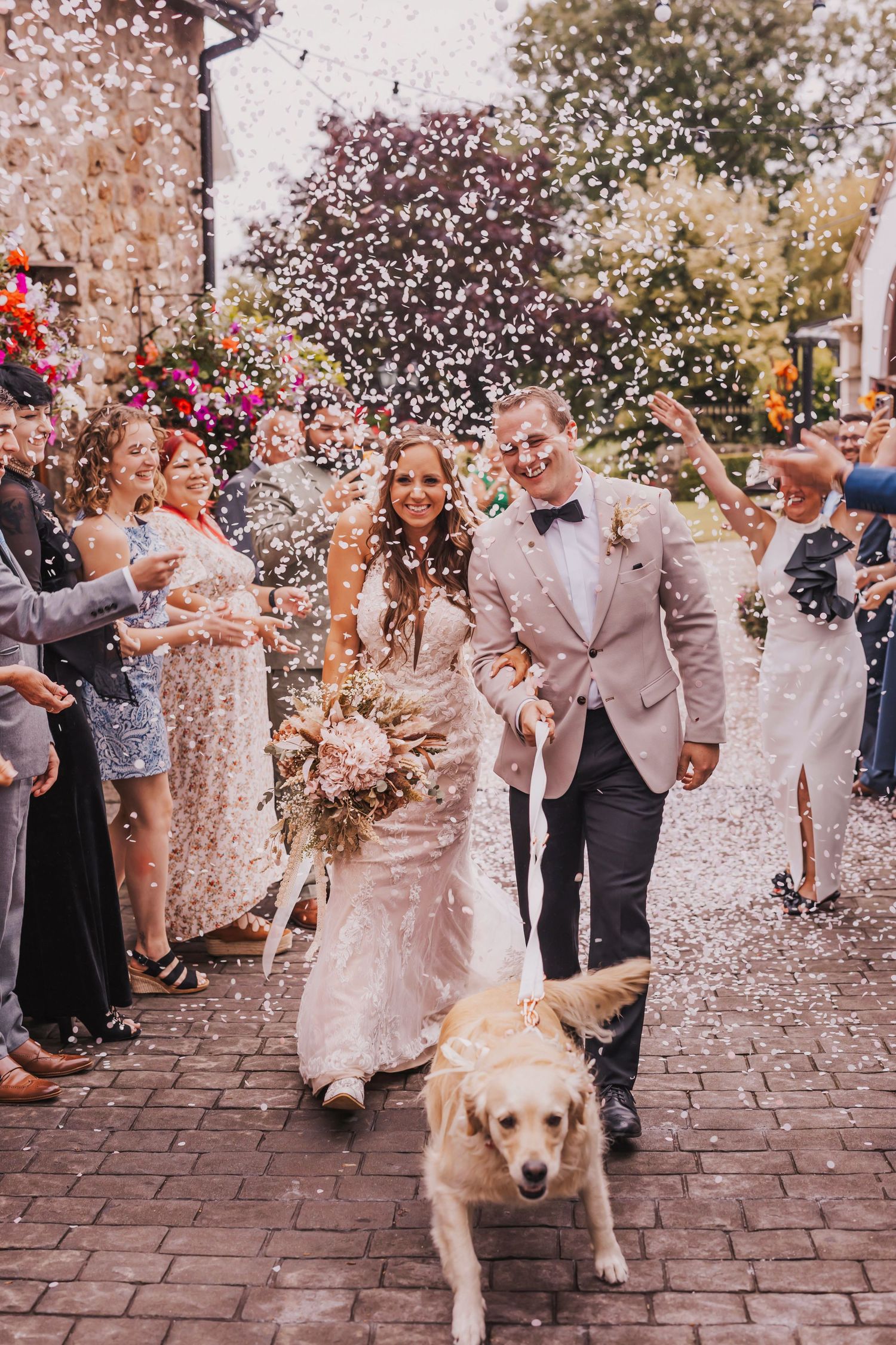 Wedding party celebrates with confetti on cobblestone path as golden retriever joins joyful recessional.