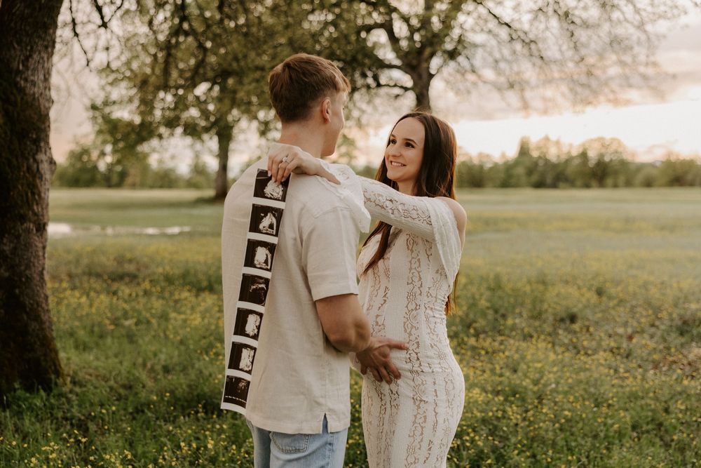 A couple in casual attire holds ultrasound photos while embracing in a sunlit meadow during golden hour.