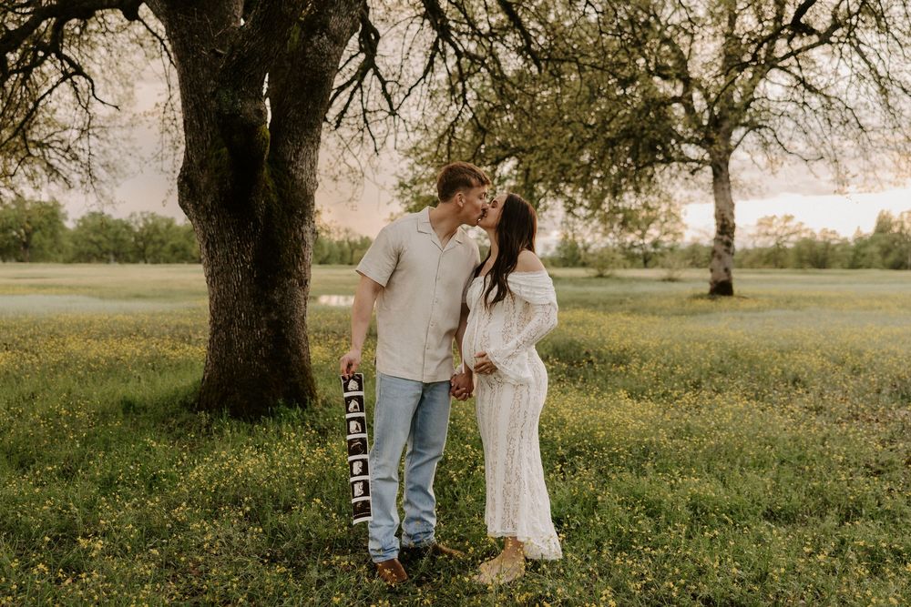 A romantic couple walks through a serene meadow with tall oak trees during golden hour sunset.