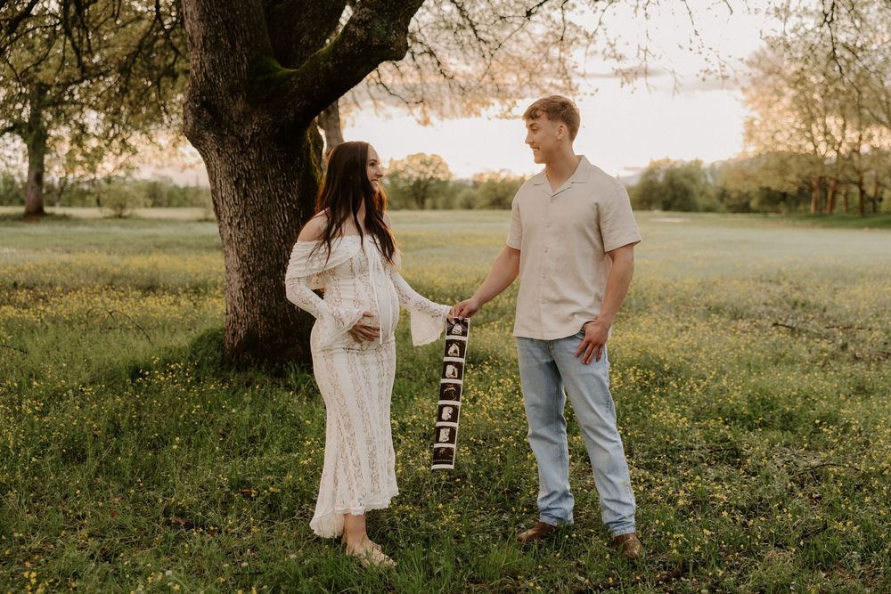A couple holds hands under a tree in a sunlit field while displaying a string of ultrasound photos.