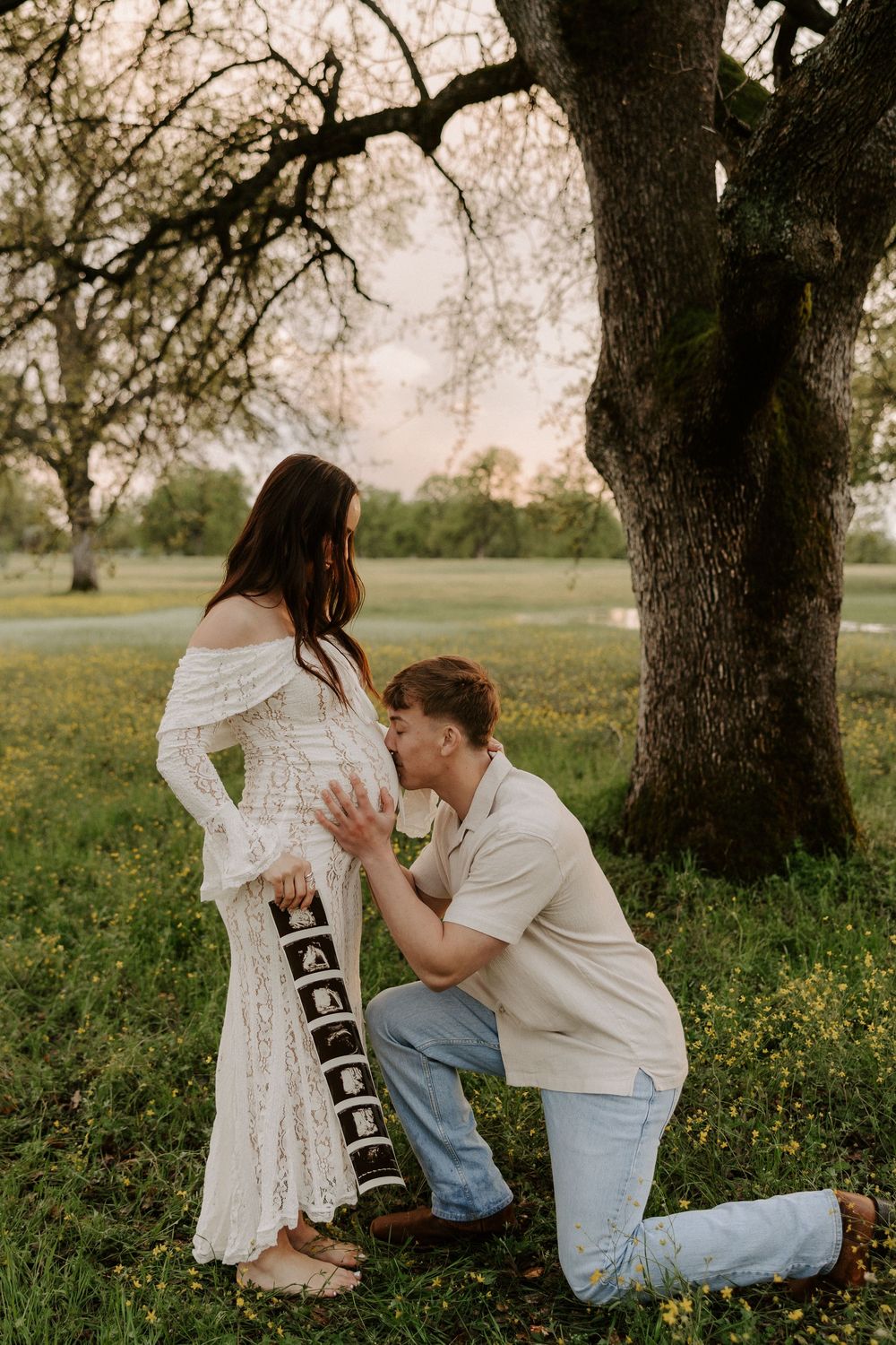 A romantic proposal scene unfolds under a large oak tree in a wildflower meadow at golden hour.