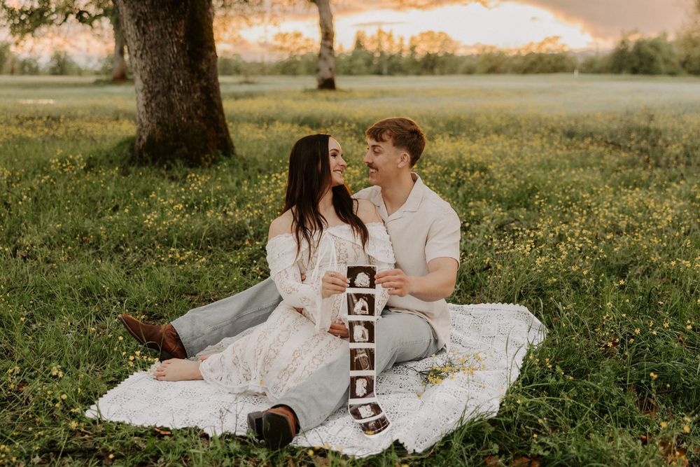 A couple in wedding attire sits on a blanket in a grassy field holding ultrasound photos at sunset.