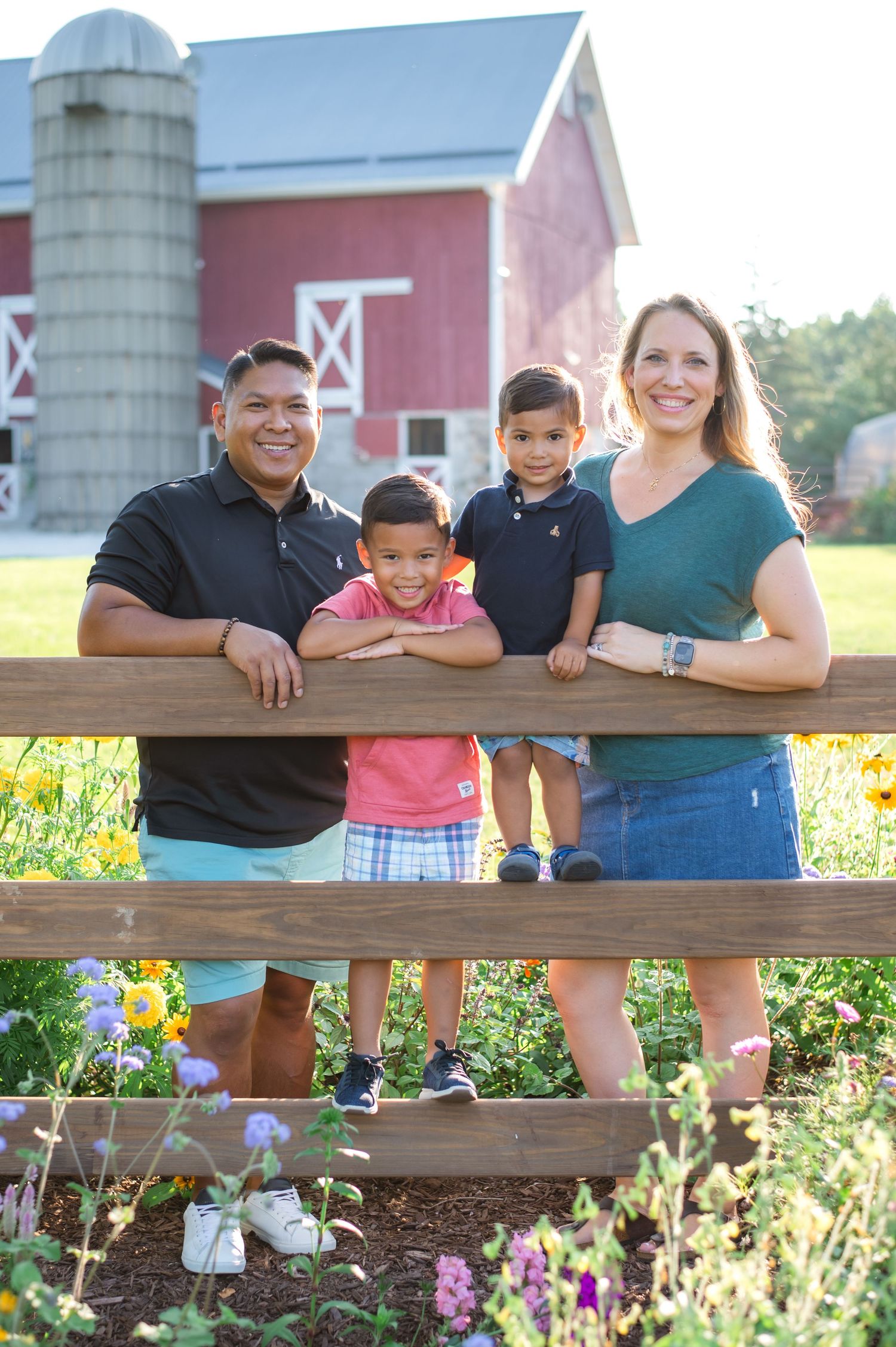 A family poses together on a wooden fence at a farm with a red barn and silo in the background.