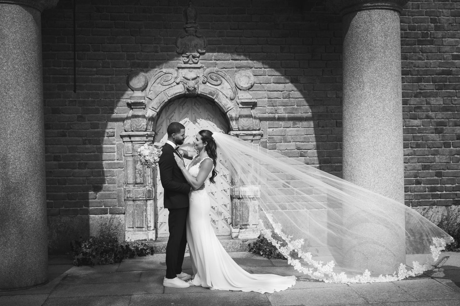 A romantic black and white wedding photo with a couple embracing under a stone archway as a long veil flows behind them.
