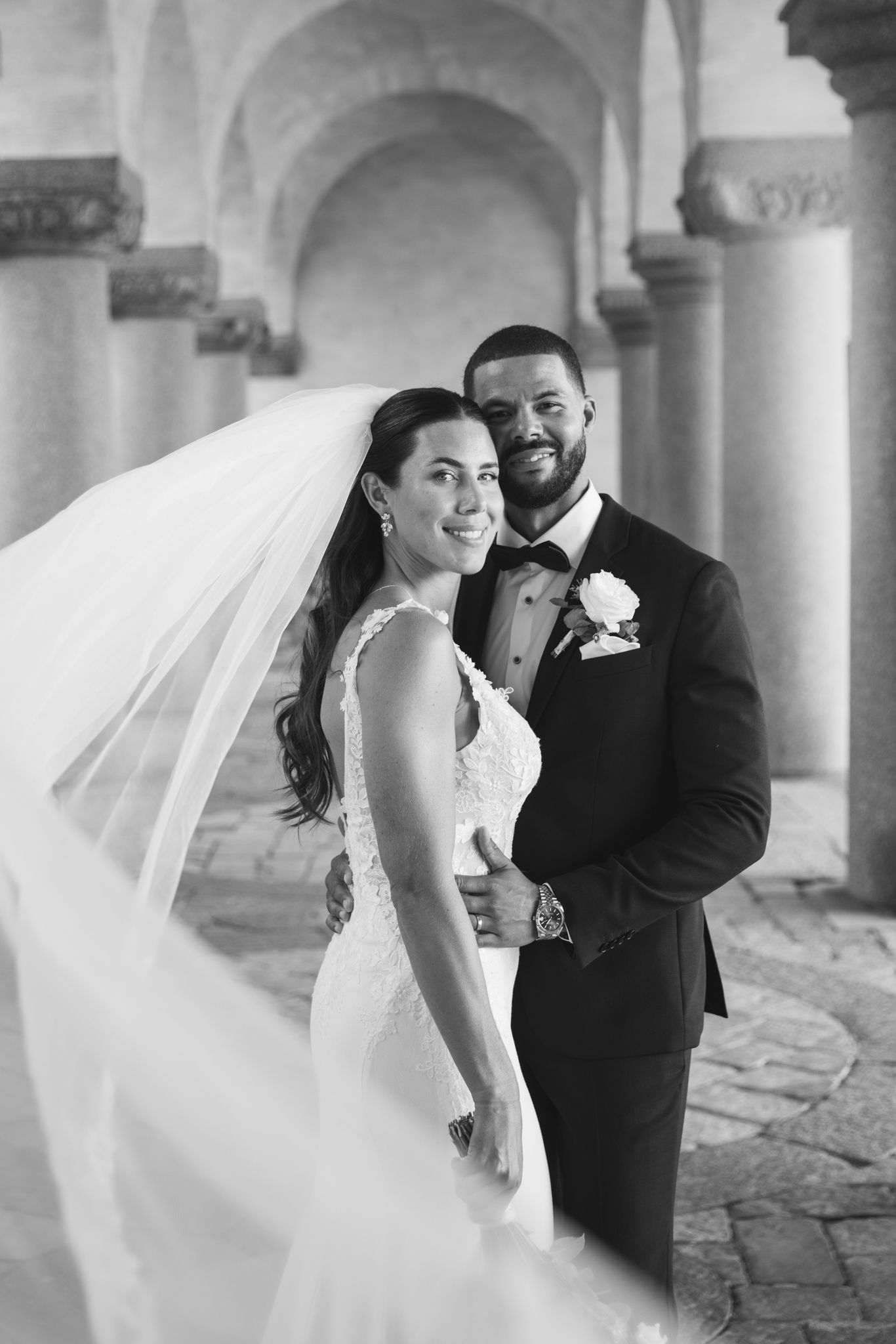A romantic black and white wedding photo series showing a couple embracing under stone archways as a veil flows in the breeze.