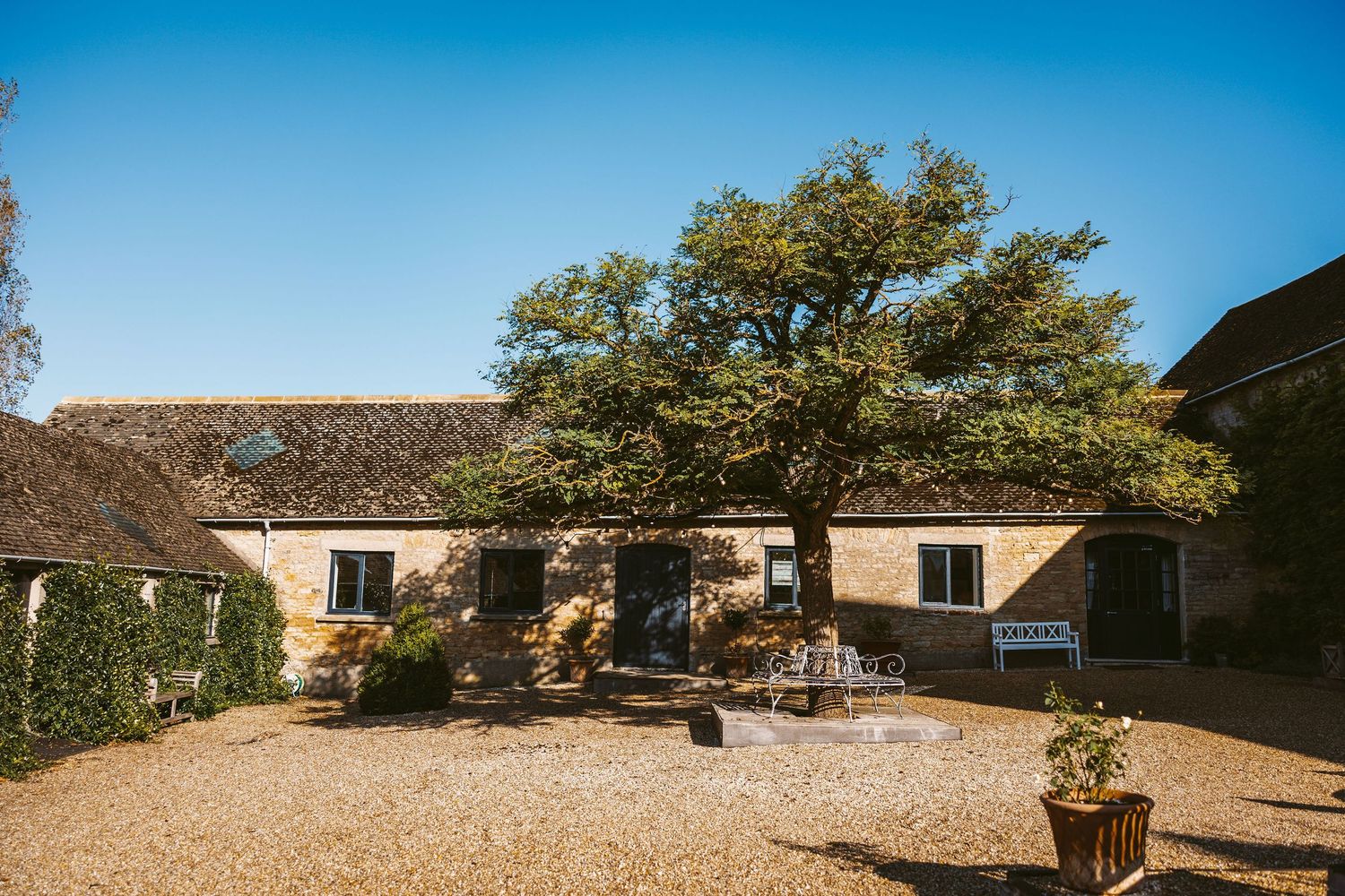 A panoramic view of a rustic stone cottage with gravel driveway and surrounding gardens under a bright blue sky.