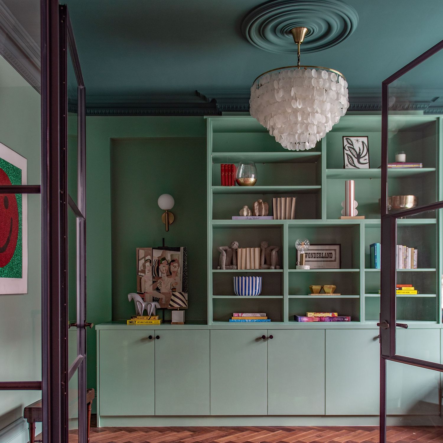 Modern home study with sage green built-in bookshelves  and an elegant capiz shell chandelier in a mint-toned room. House of Hackney paints