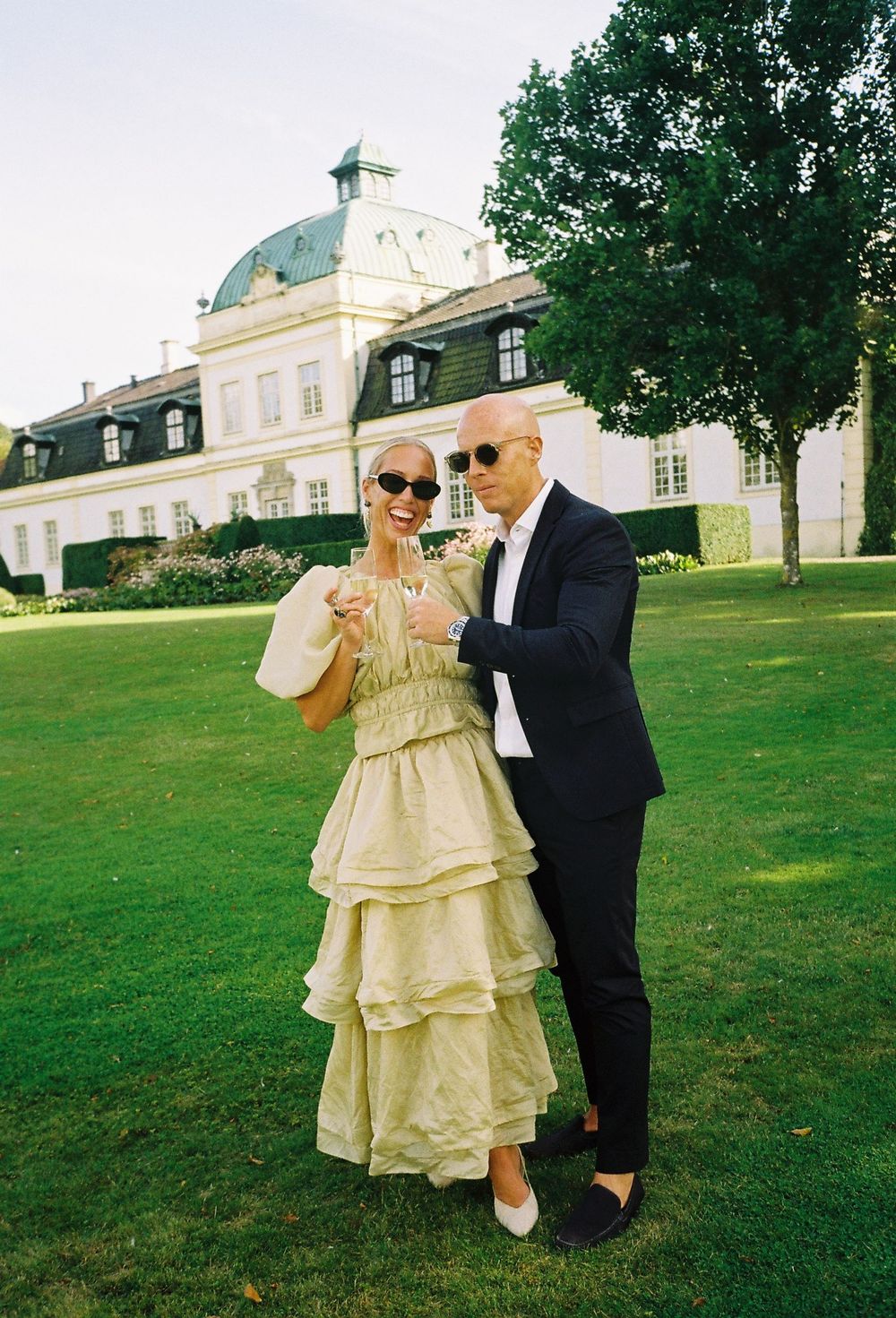 Couple in elegant attire posing on green lawn before grand white manor house with dome roof.