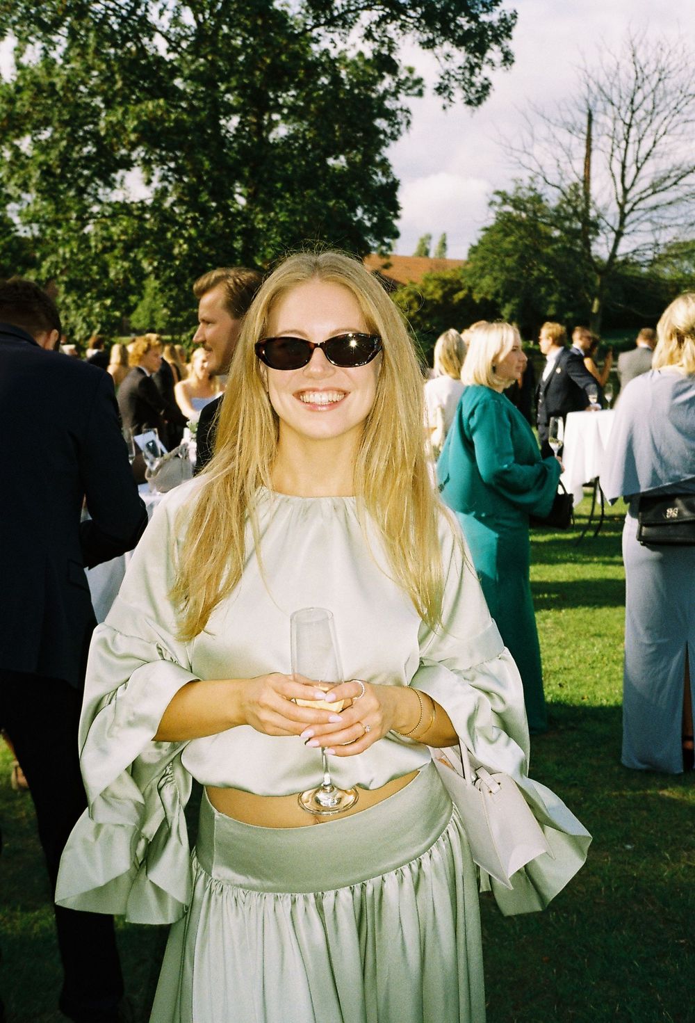 Smiling blonde woman in mint green outfit with sunglasses holding champagne flute at outdoor garden party.