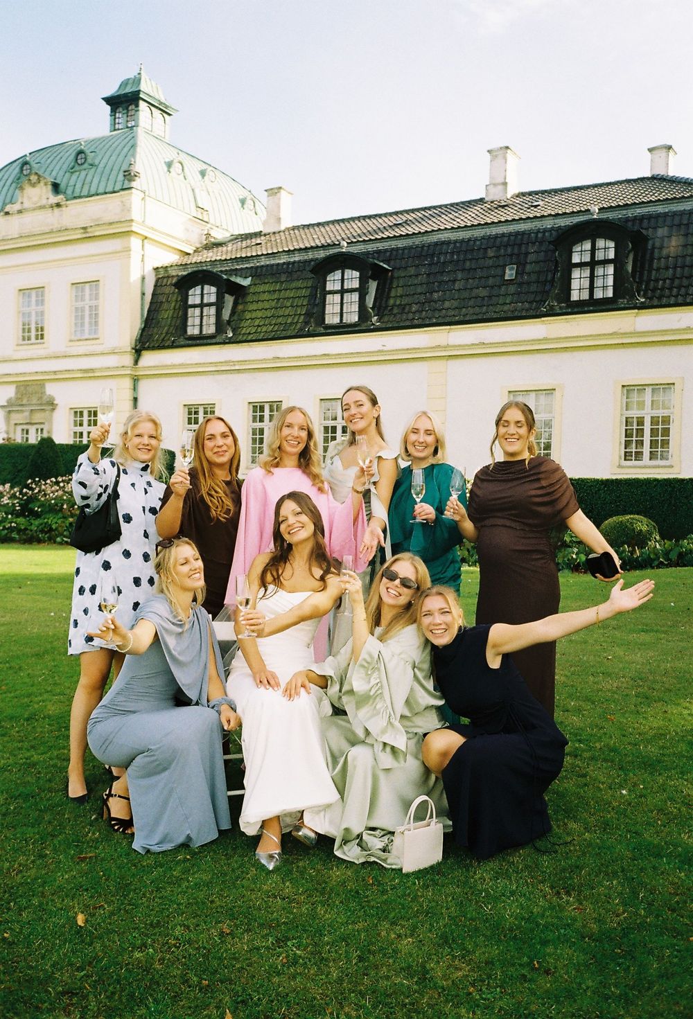 Group of women celebrating outdoors at elegant white manor house, holding champagne glasses, dressed in colorful formal attire.