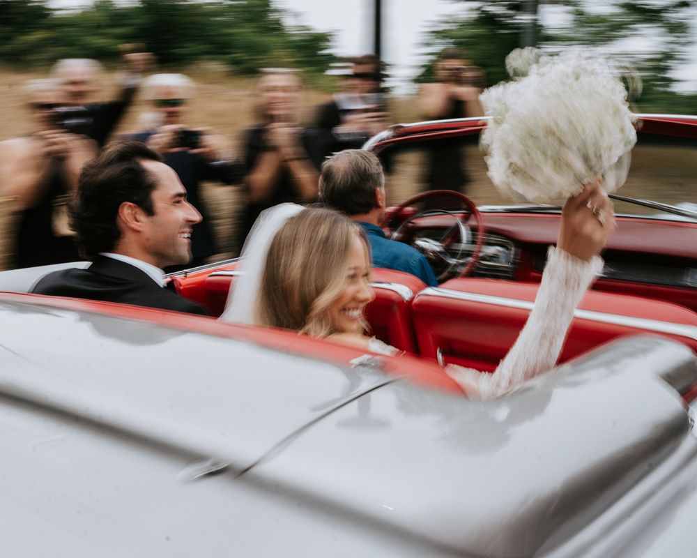 Bride and groom riding in a red convertible classic car, bride waving bouquet joyfully on their wedding day.