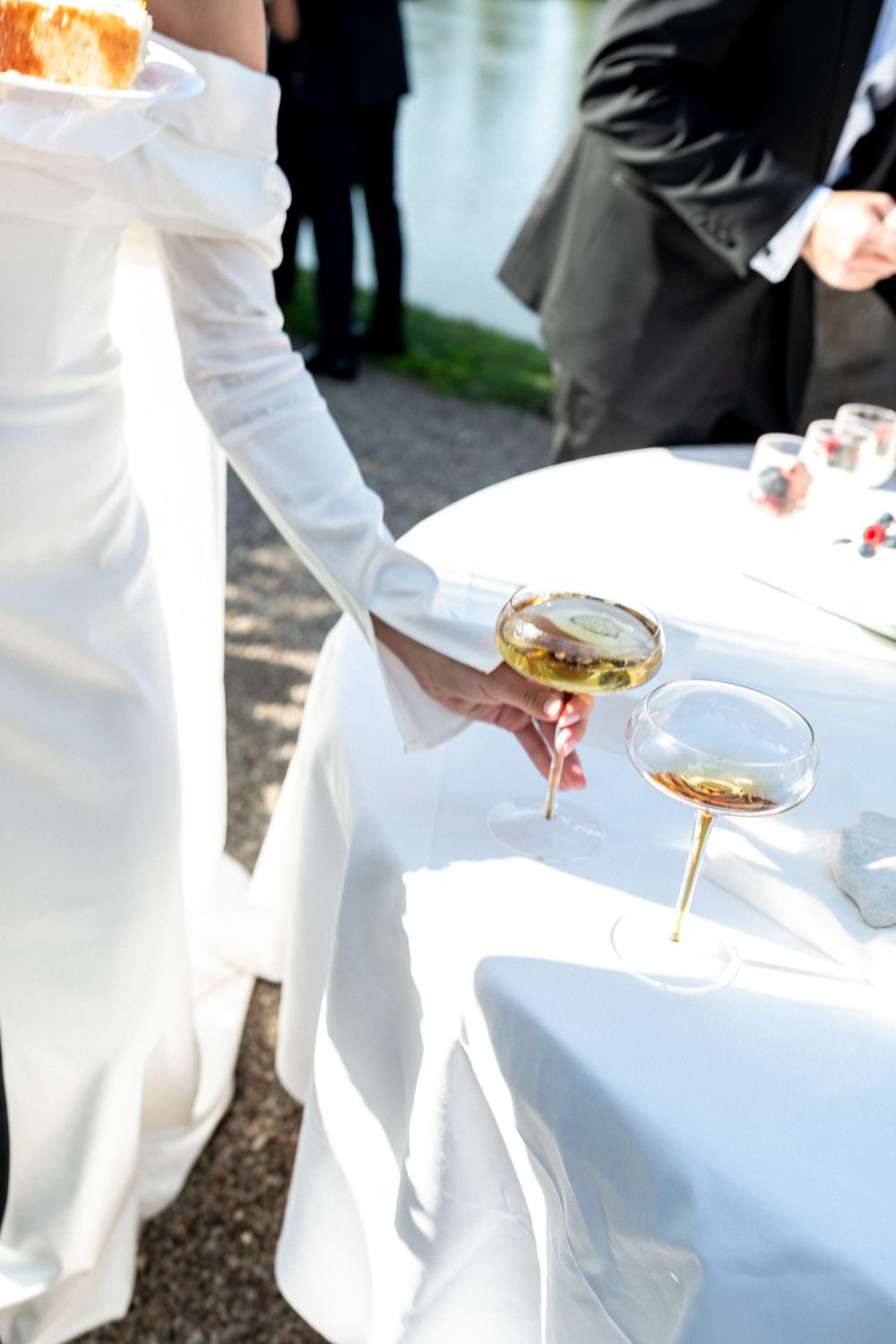 Bride in white gown holding champagne coupe glass at outdoor wedding reception table with wine glasses.