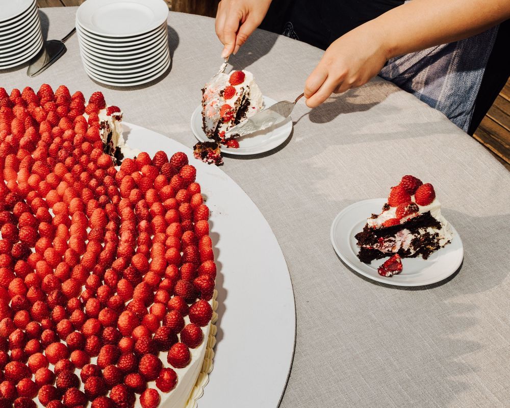 Person serving a slice of large strawberry-topped cake onto a white plate at a table.