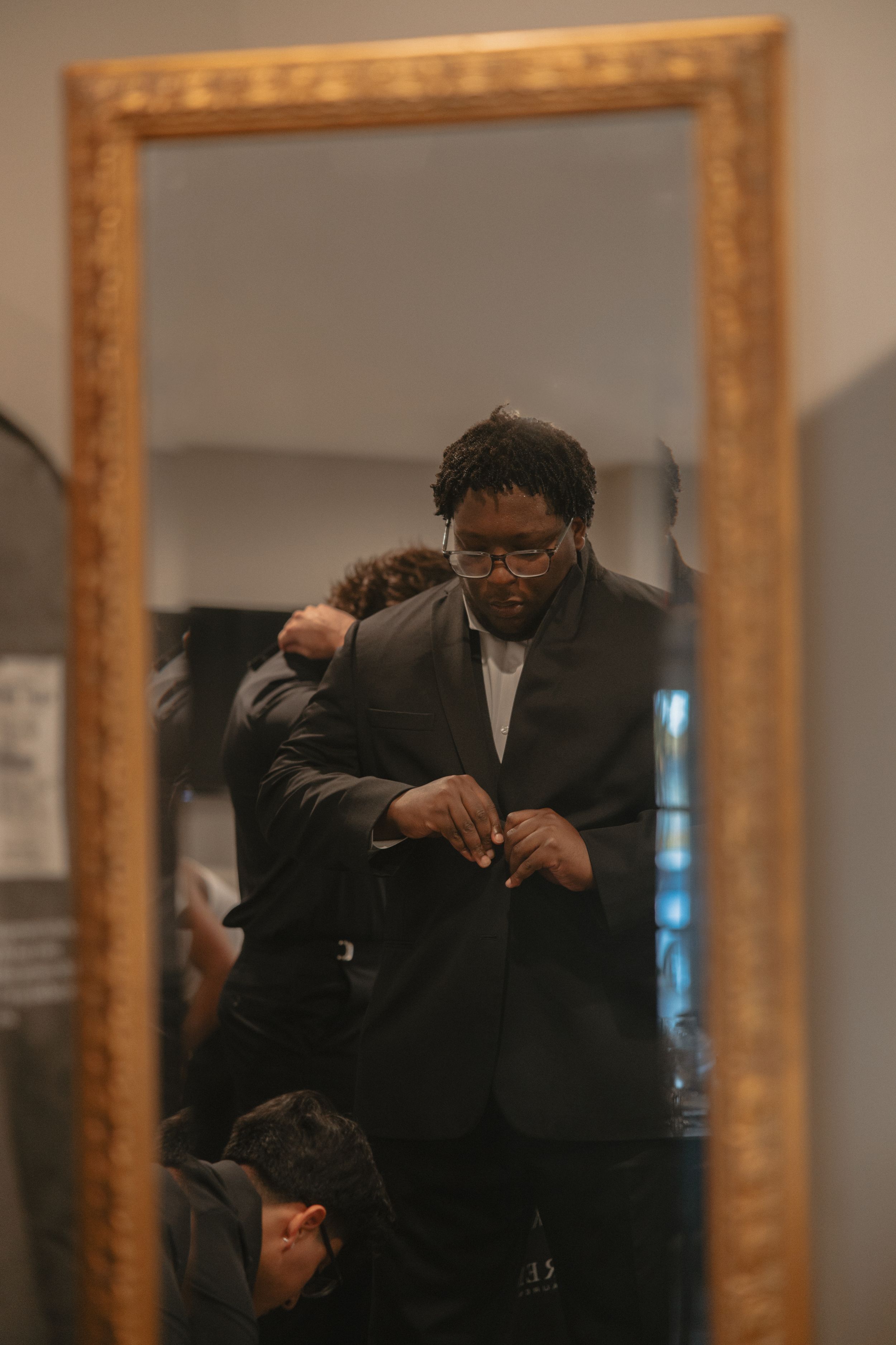 A person in a black suit adjusts their tie while looking in a series of gold-framed mirrors.