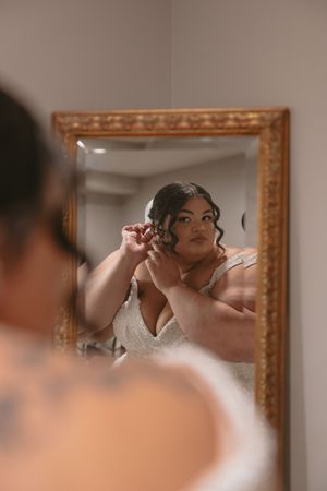 A bride putting on earrings while looking in an ornate gold-framed mirror during wedding preparations.